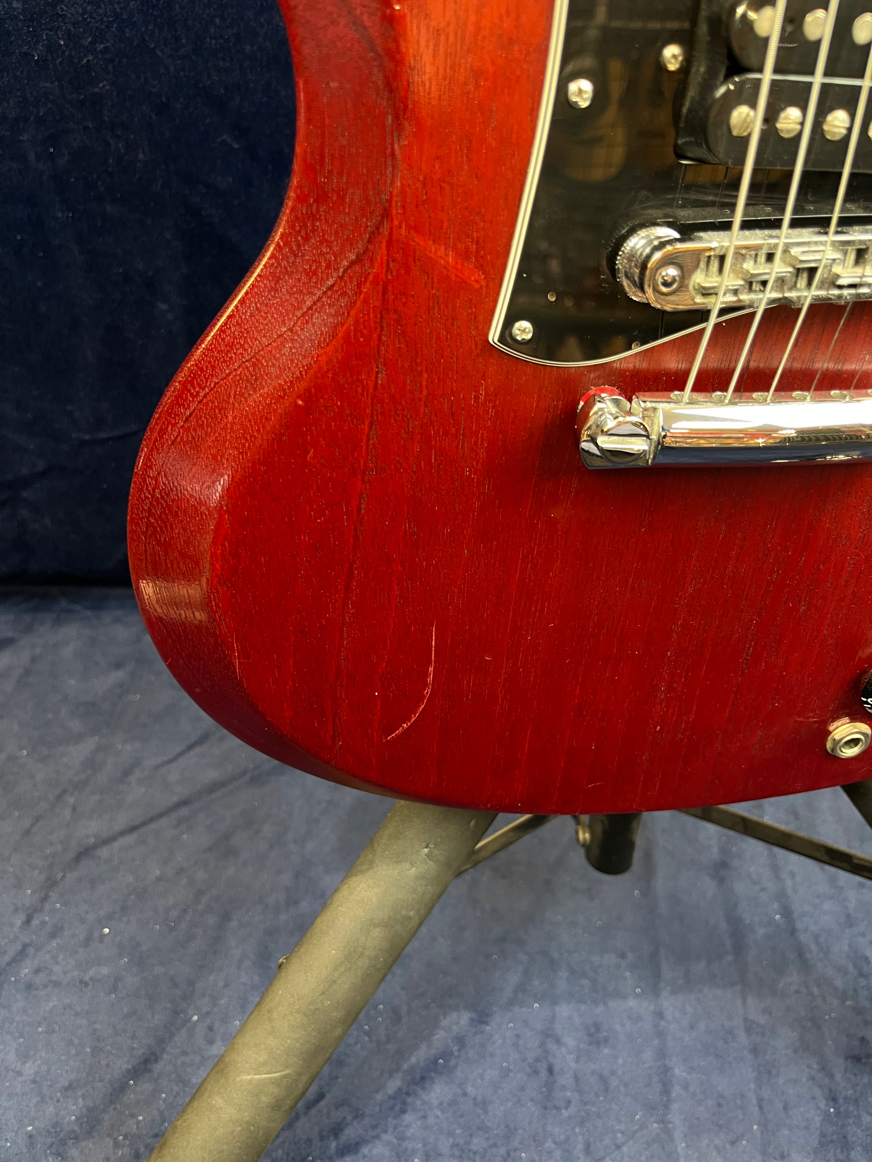 Red electric guitar body with a visible scratch near the bottom edge, chrome bridge, black pickguard, and part of the string area in view.
