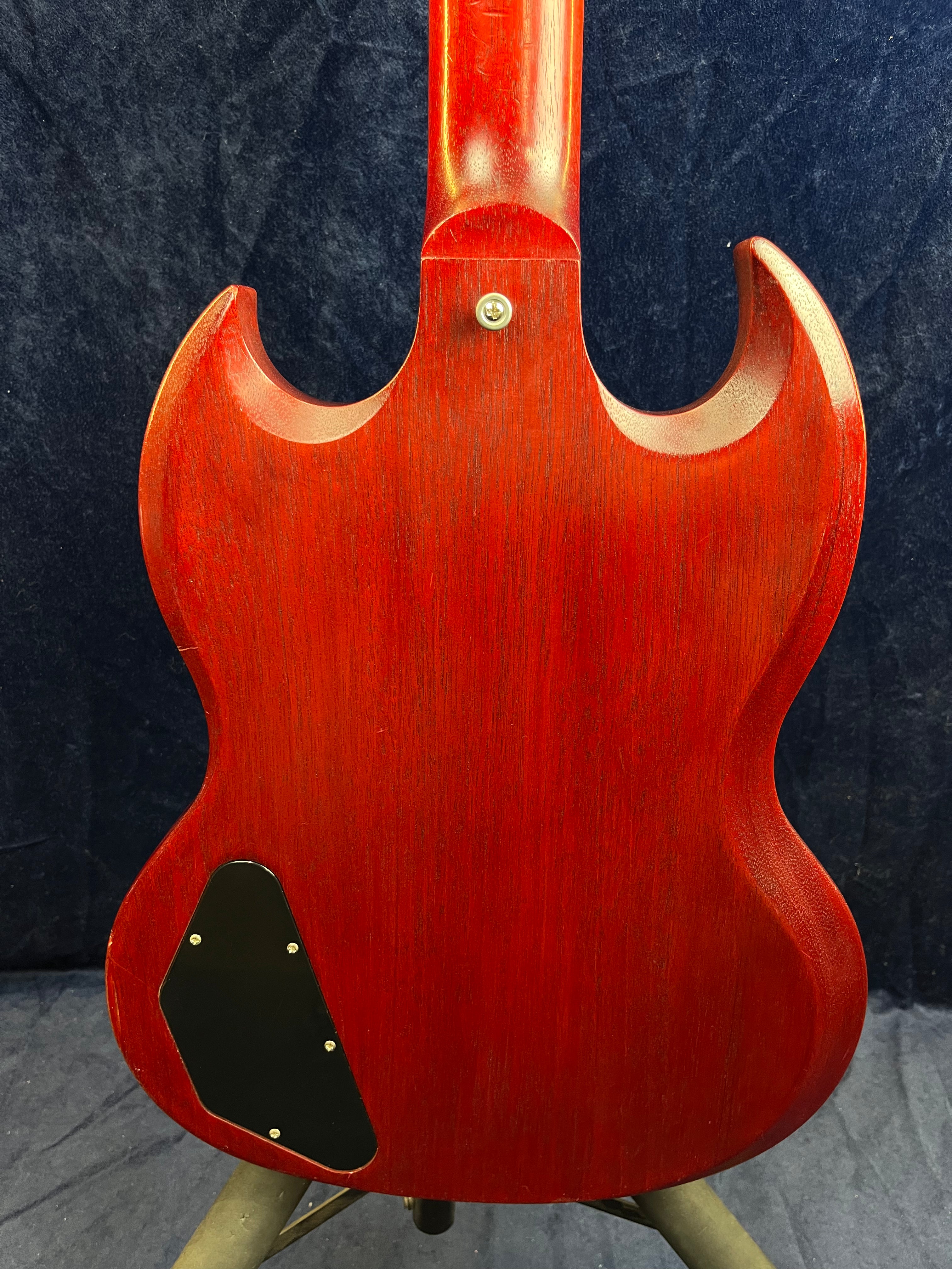 Back of a red electric guitar with a double cutaway body, black electronics cover plate, and visible wood grain against a dark background.