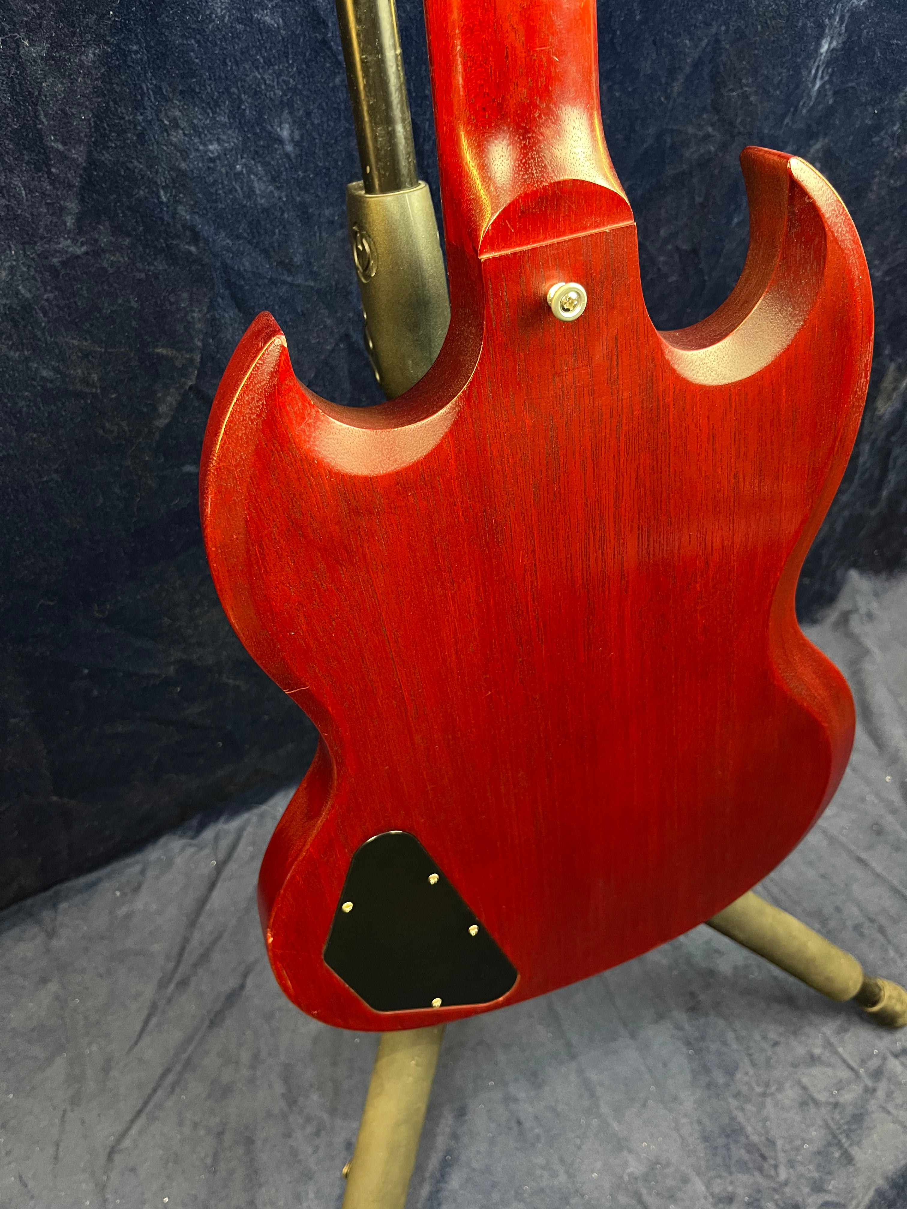 Back of a red electric guitar with double cutaway design, black triangular electronics cover, and a guitar stand visible at the neck joint.