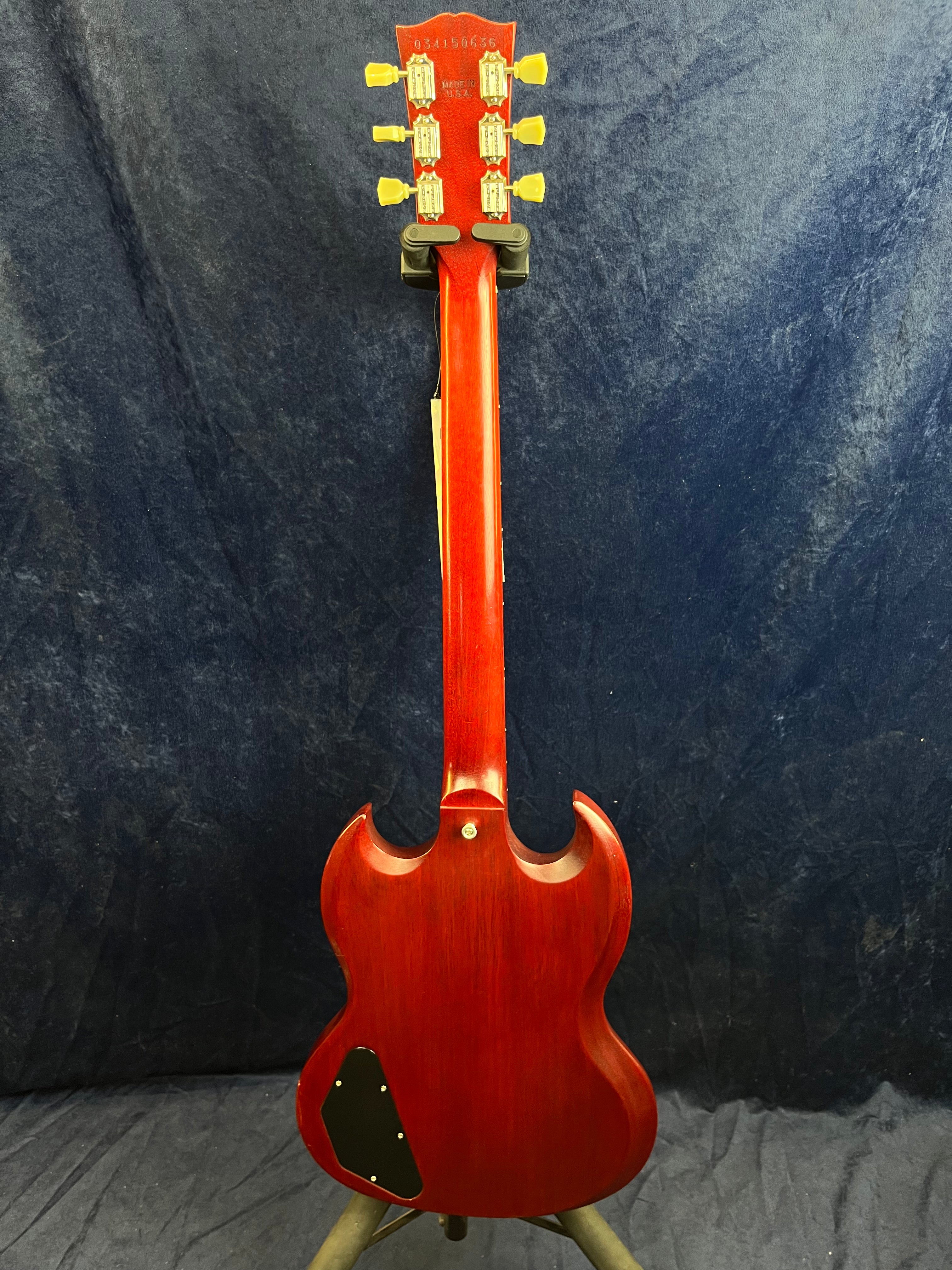 A red electric guitar with a double-cutaway body and yellow tuning pegs is shown from the back, resting on a stand against a dark background.