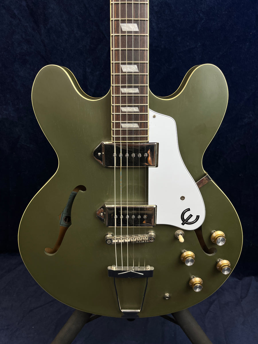 Olive green semi-hollow electric guitar with white pickguard, two pickups, four gold control knobs, and decorative f-holes on the body.