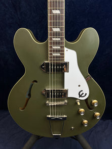 Olive green semi-hollow electric guitar with white pickguard, two pickups, four gold control knobs, and decorative f-holes on the body.