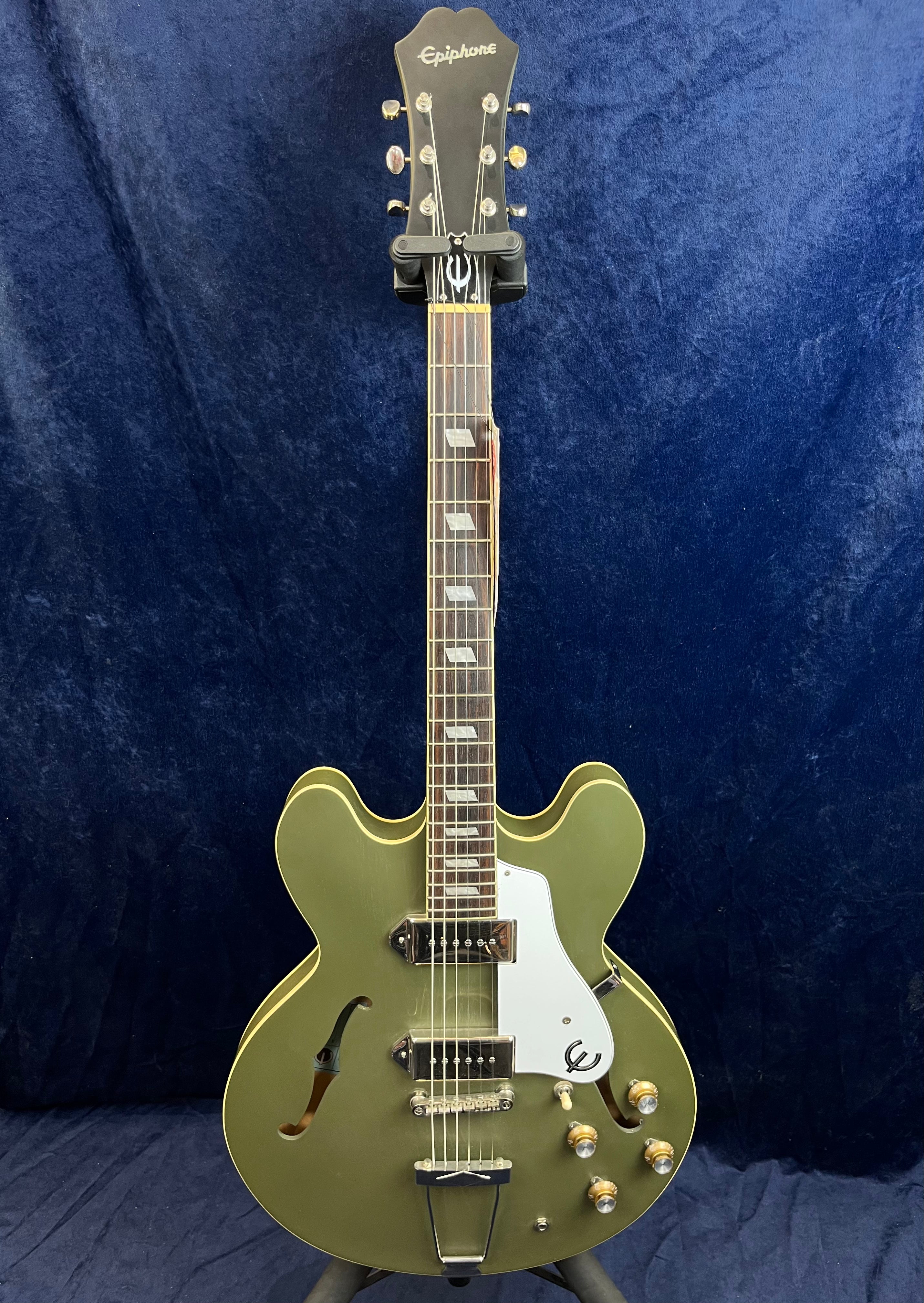 Olive green Epiphone semi-hollow electric guitar with two pickups, white pickguard, gold hardware, and double cutaway body shape against a dark background.