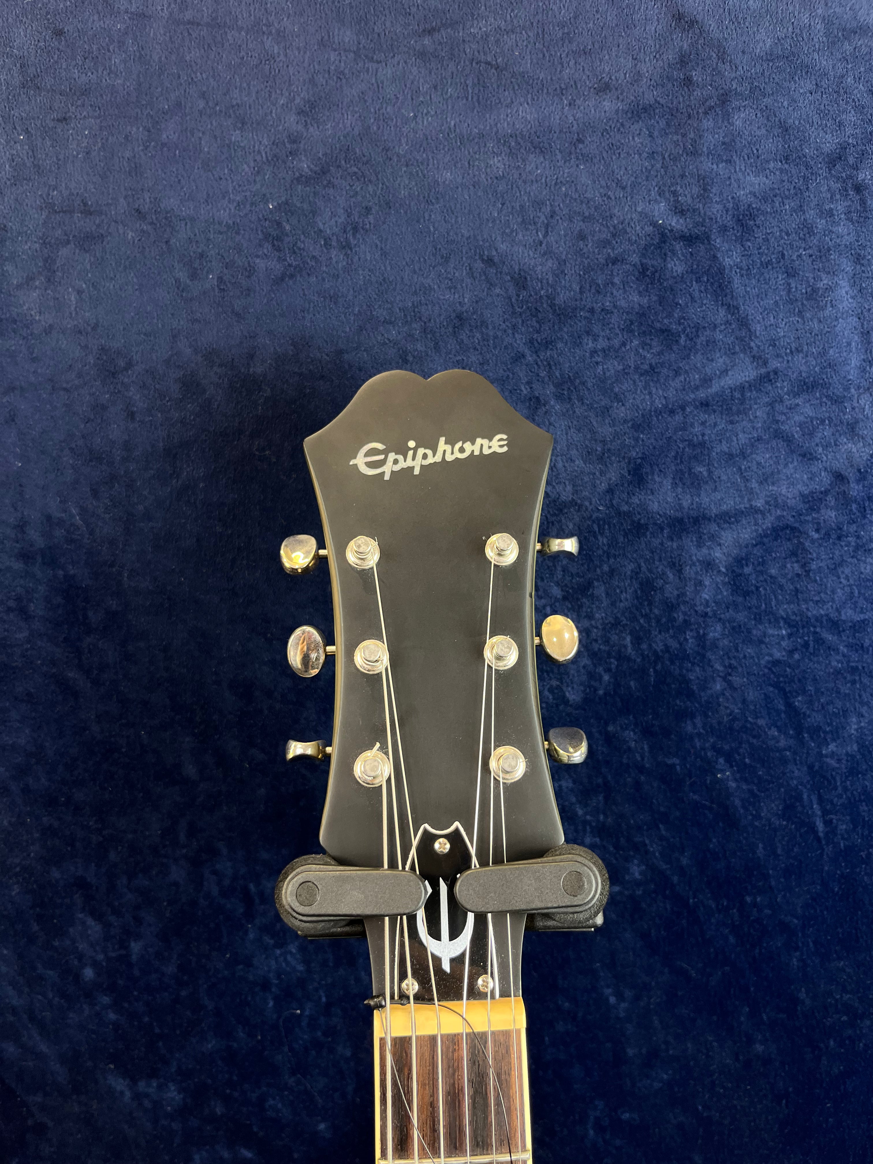 Epiphone electric guitar headstock with six tuning pegs, black finish, silver logo, and a dark wood fretboard against a dark blue background.