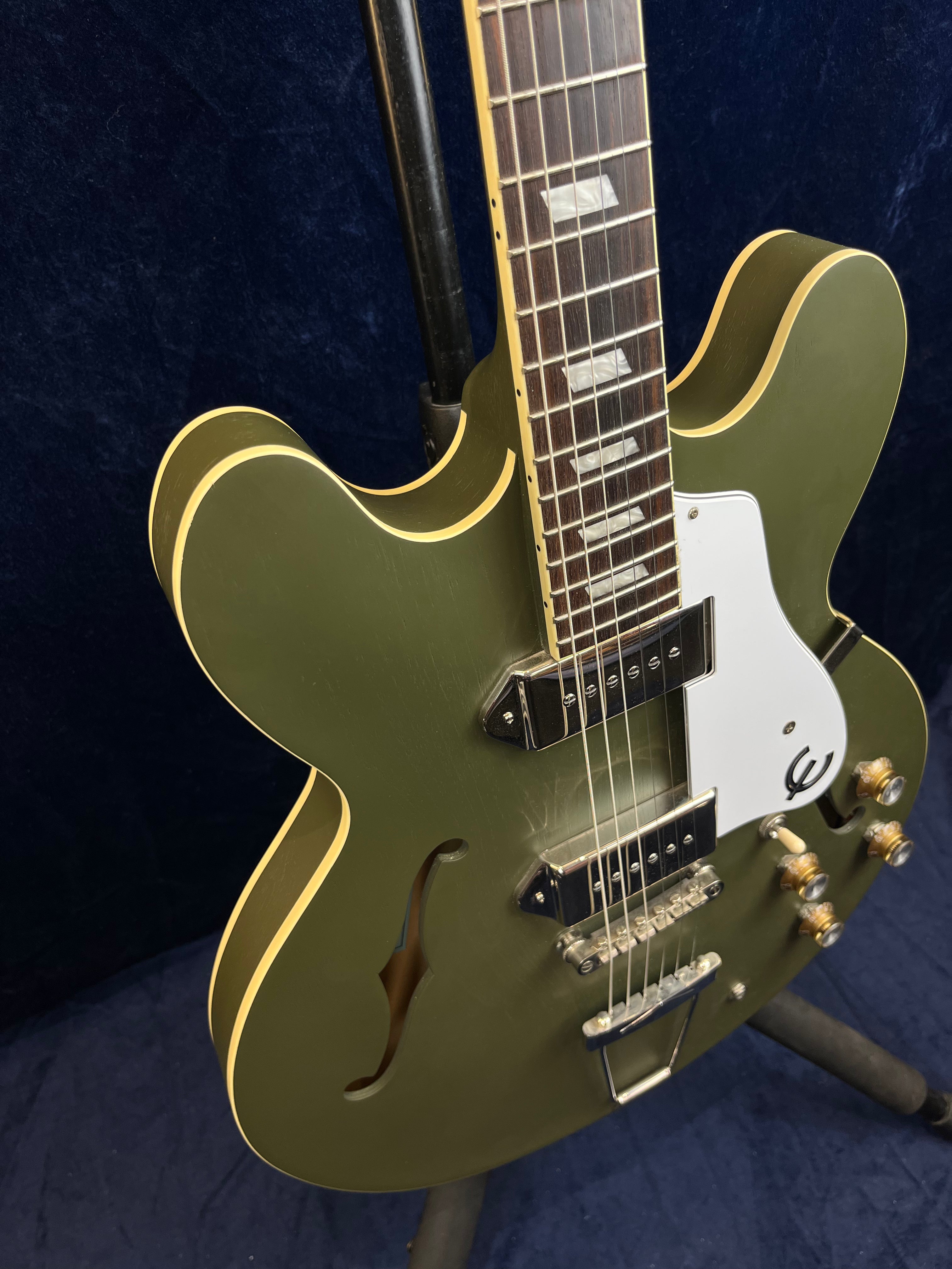 Olive green semi-hollow electric guitar with cream binding, white pickguard, and chrome hardware, featuring dual pickups and four amber control knobs.