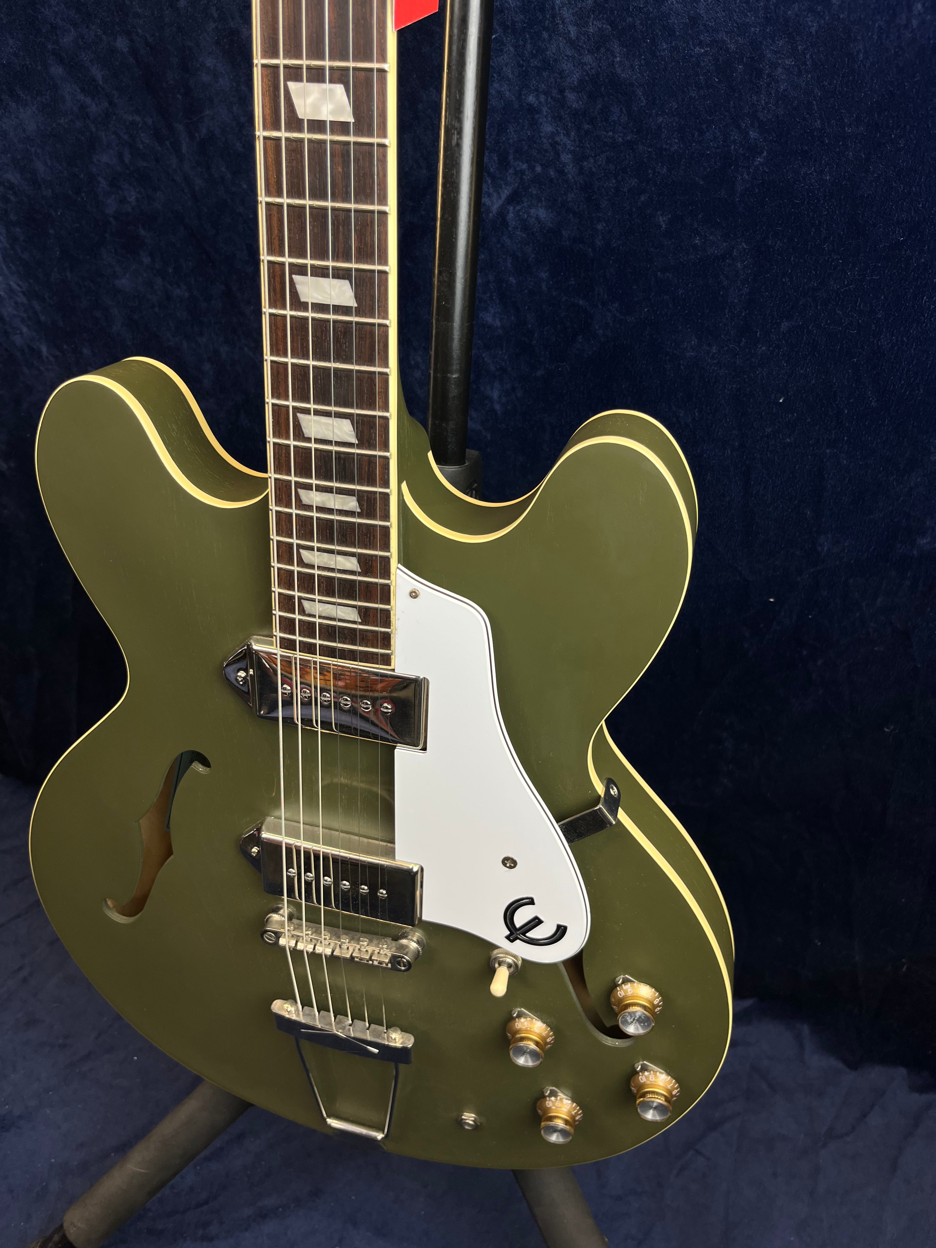 Olive green semi-hollow body electric guitar with white pickguard, two pickups, gold control knobs, and cream binding, displayed on a stand.