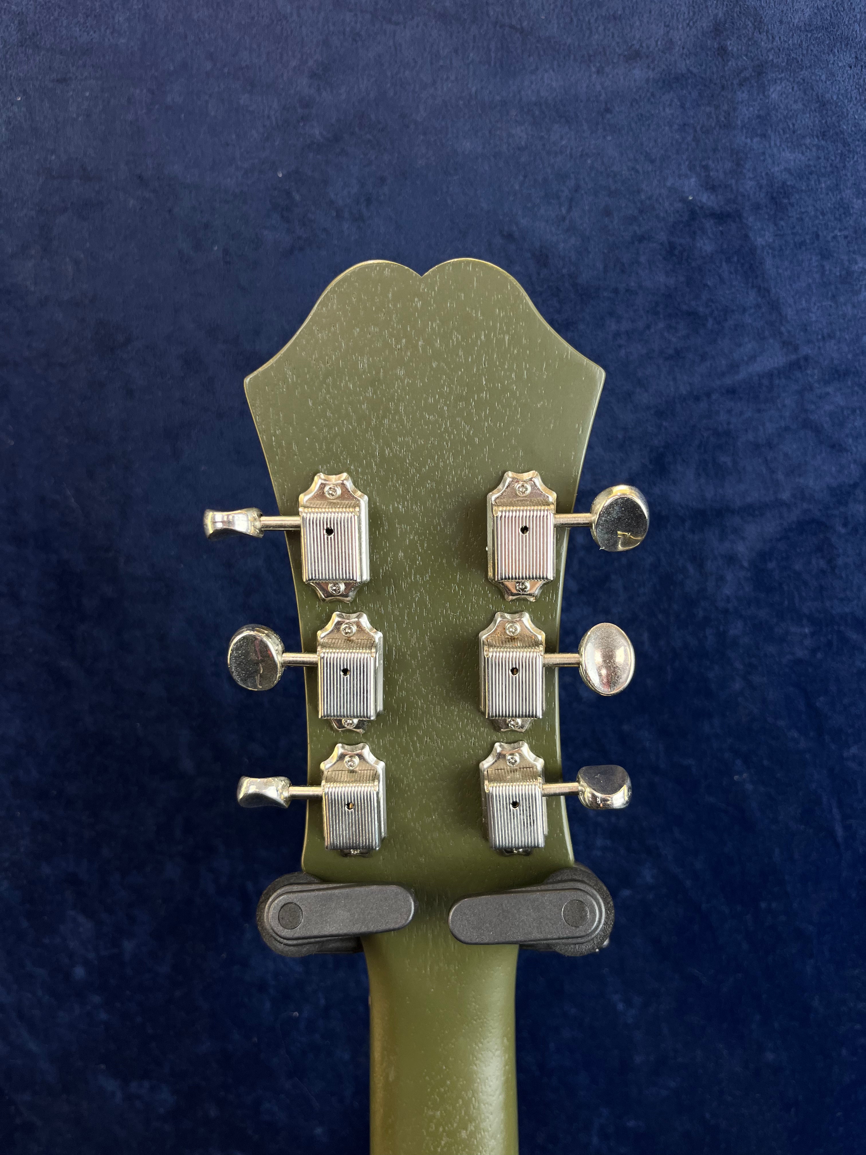 Olive green electric guitar headstock with six vintage-style silver tuning pegs, photographed against a dark blue textured background.