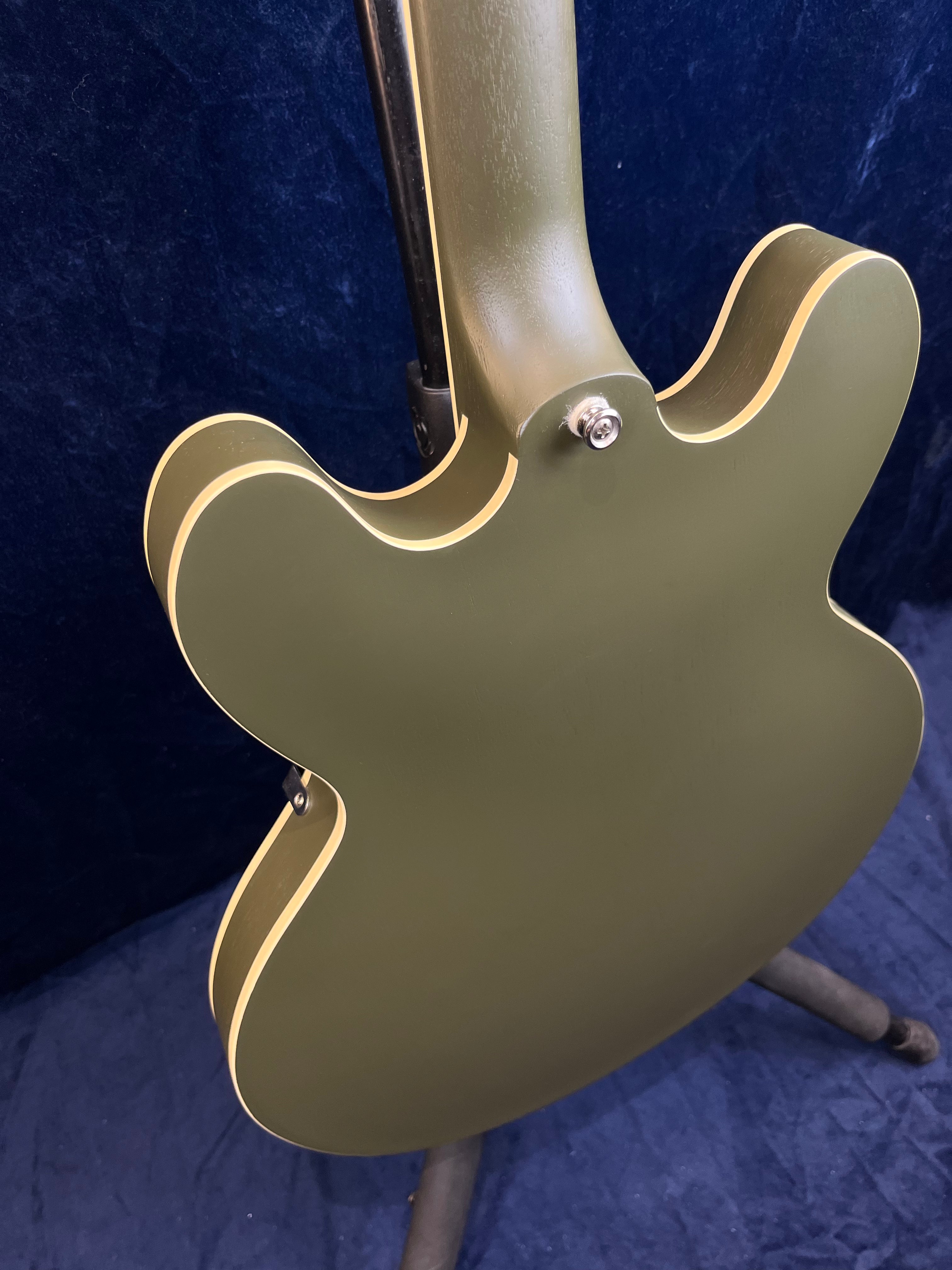 Olive green semi-hollow electric guitar body with cream binding, viewed from the back while standing on a guitar stand.