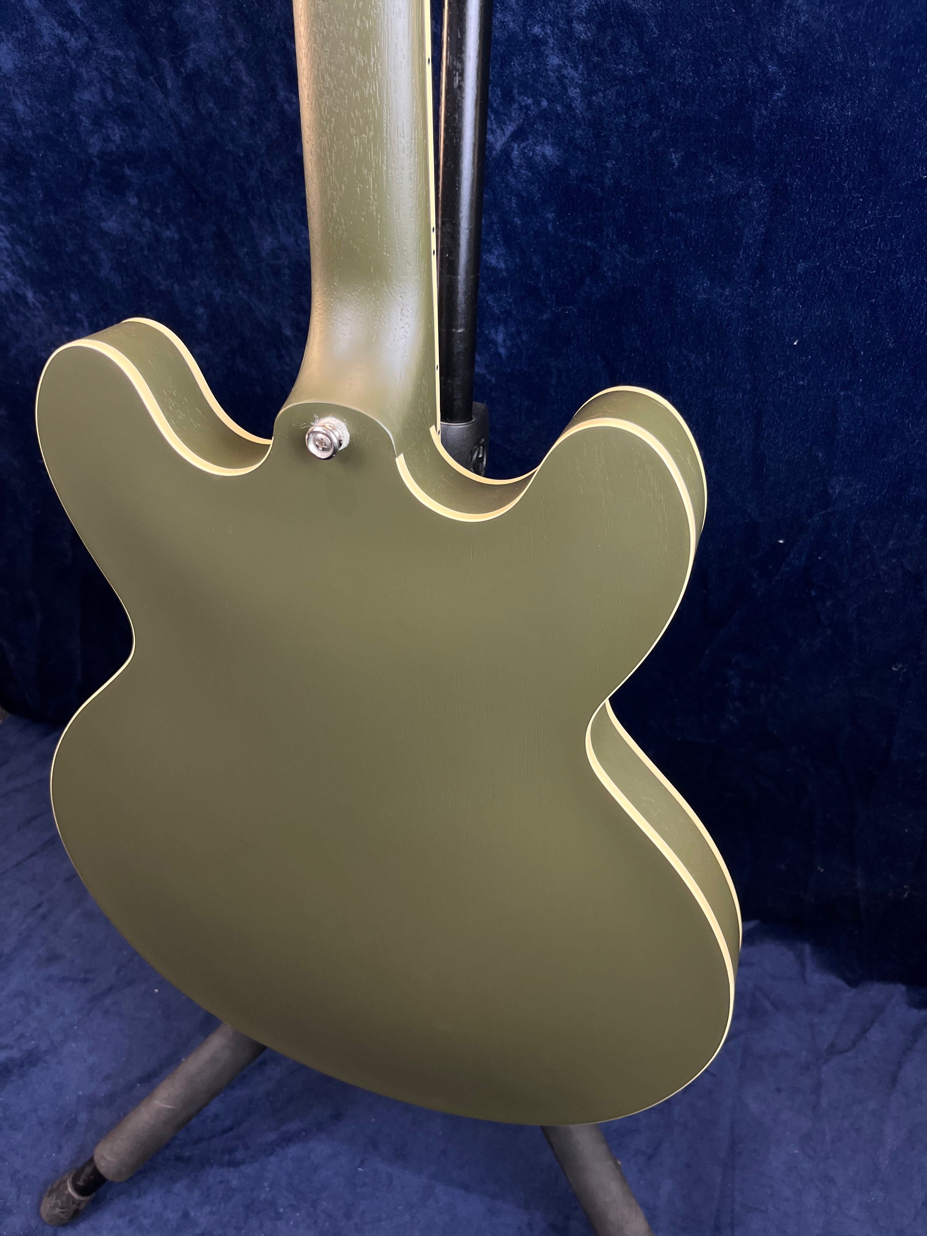 Olive green semi-hollow electric guitar viewed from the back, with cream-colored binding outlining the body and neck, resting on a stand.
