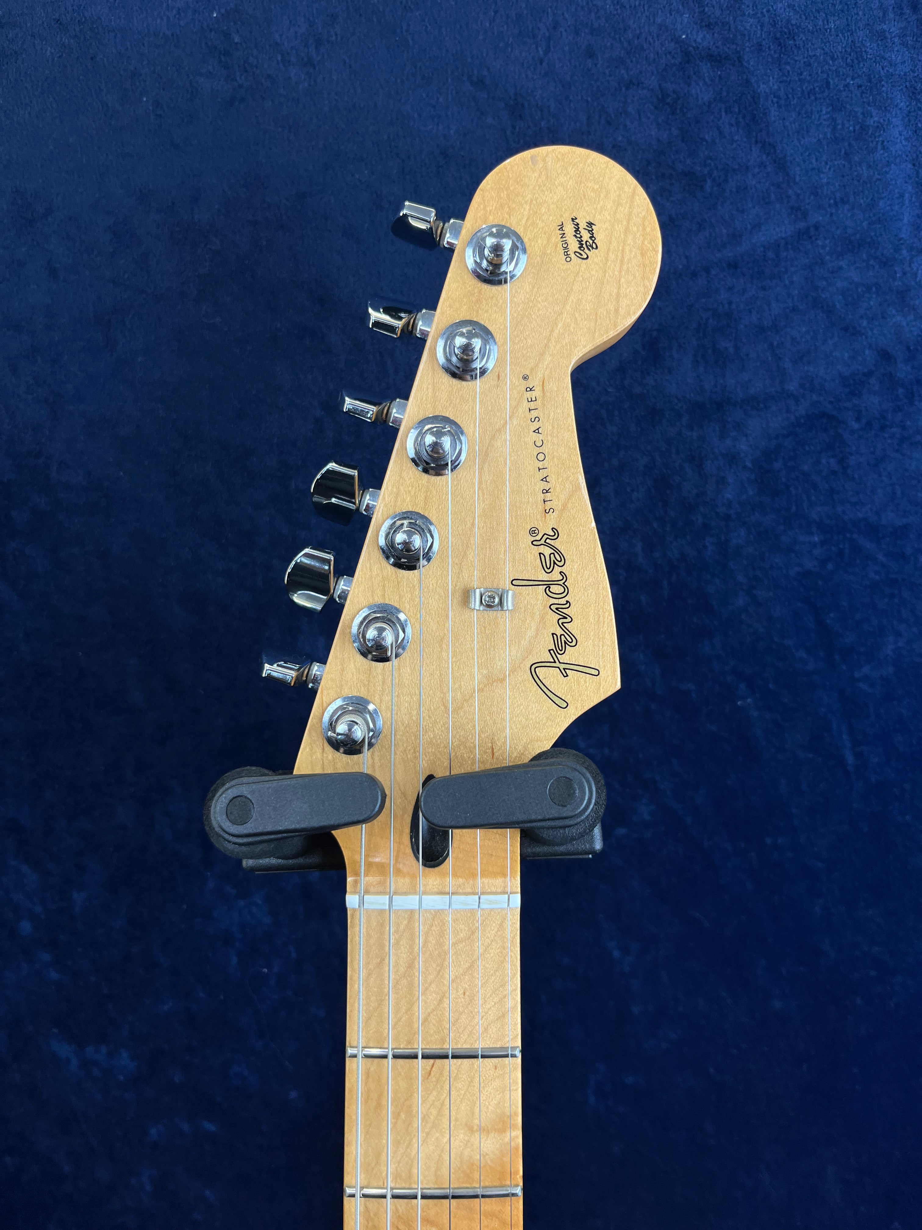 Tête d’une guitare électrique Fender Stratocaster en bois clair, six mécaniques chromées alignées, fond bleu foncé, support noir attaché au manche.