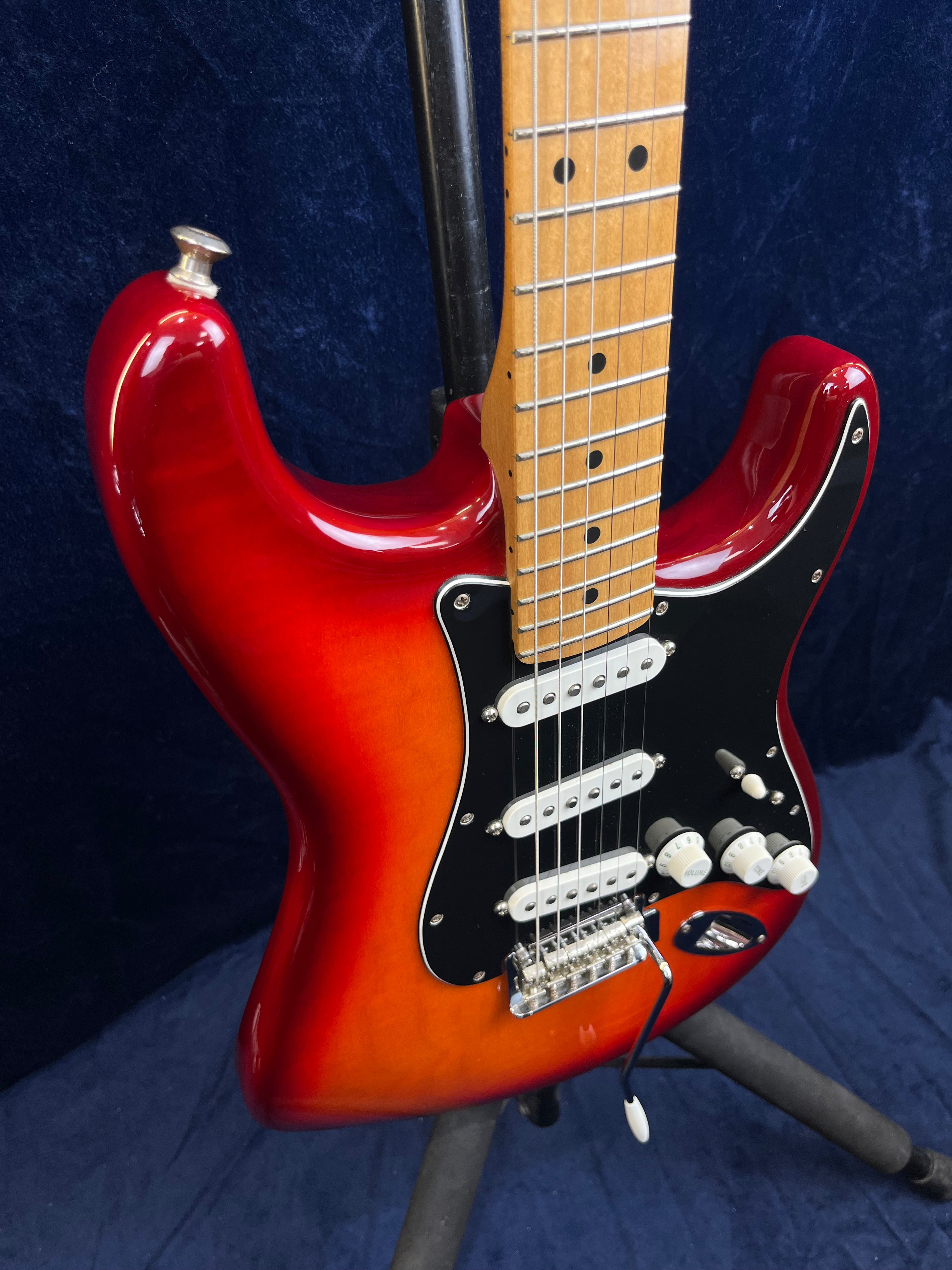 Guitare électrique rouge et orange à trois micros simples, plaque noire, boutons blancs et manche en bois clair, posée sur un support noir.