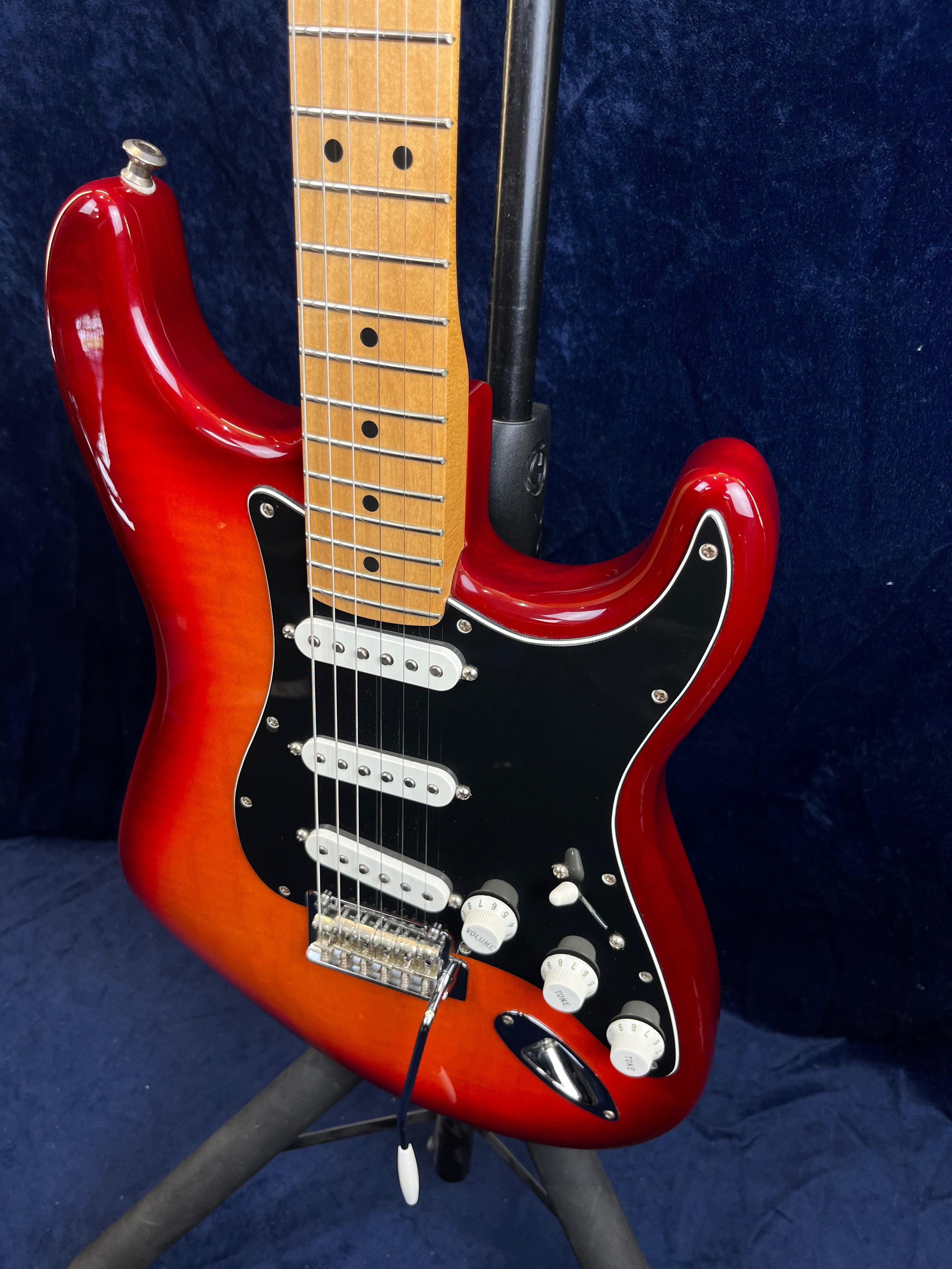 Guitare électrique rouge avec manche en érable, trois micros simples blancs et plaque de protection noire, montée sur pied.