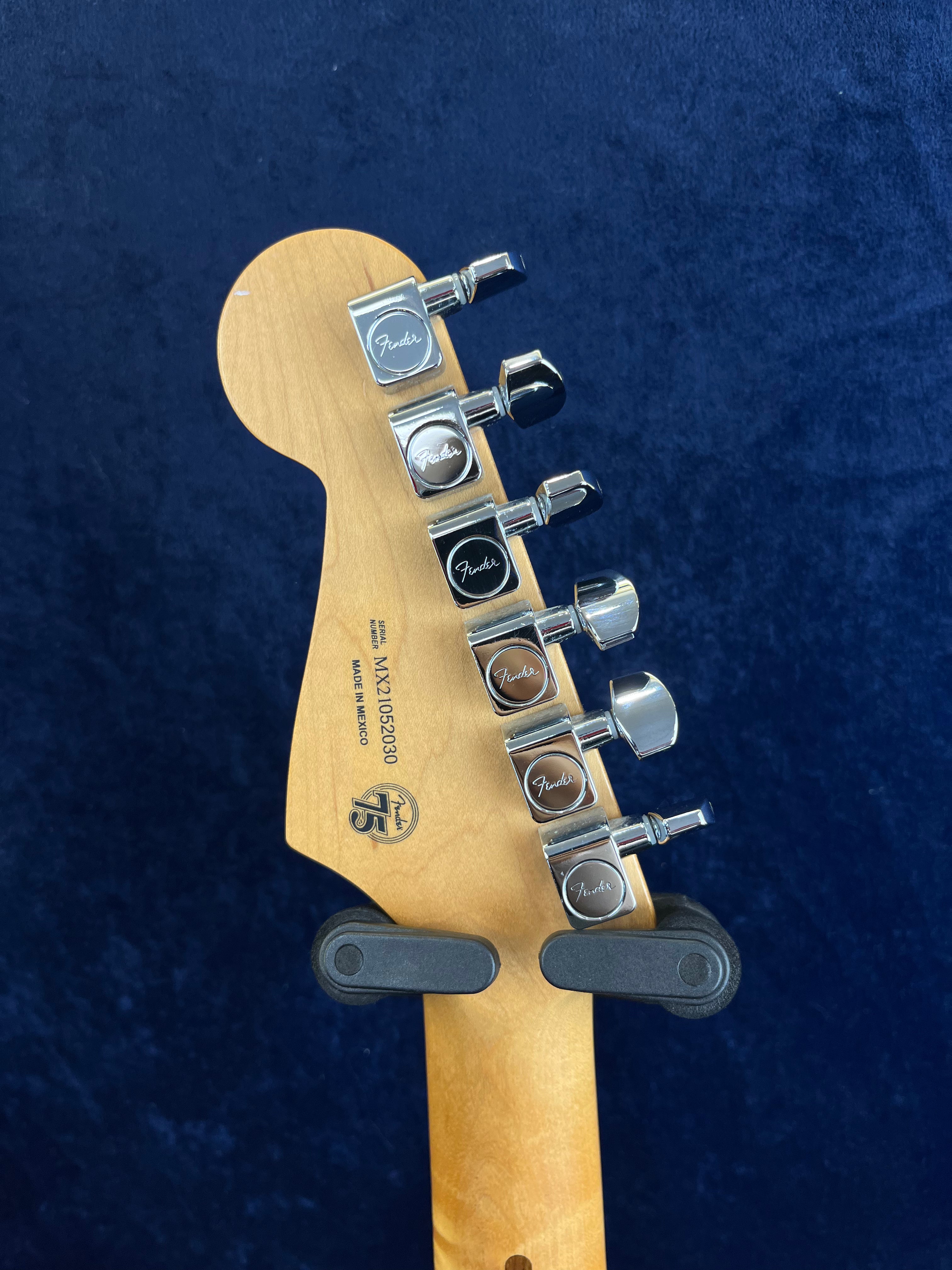 Tête d'une guitare électrique Fender en bois clair, six chevilles d’accordage chromées alignées, support mural noir, fond bleu foncé.