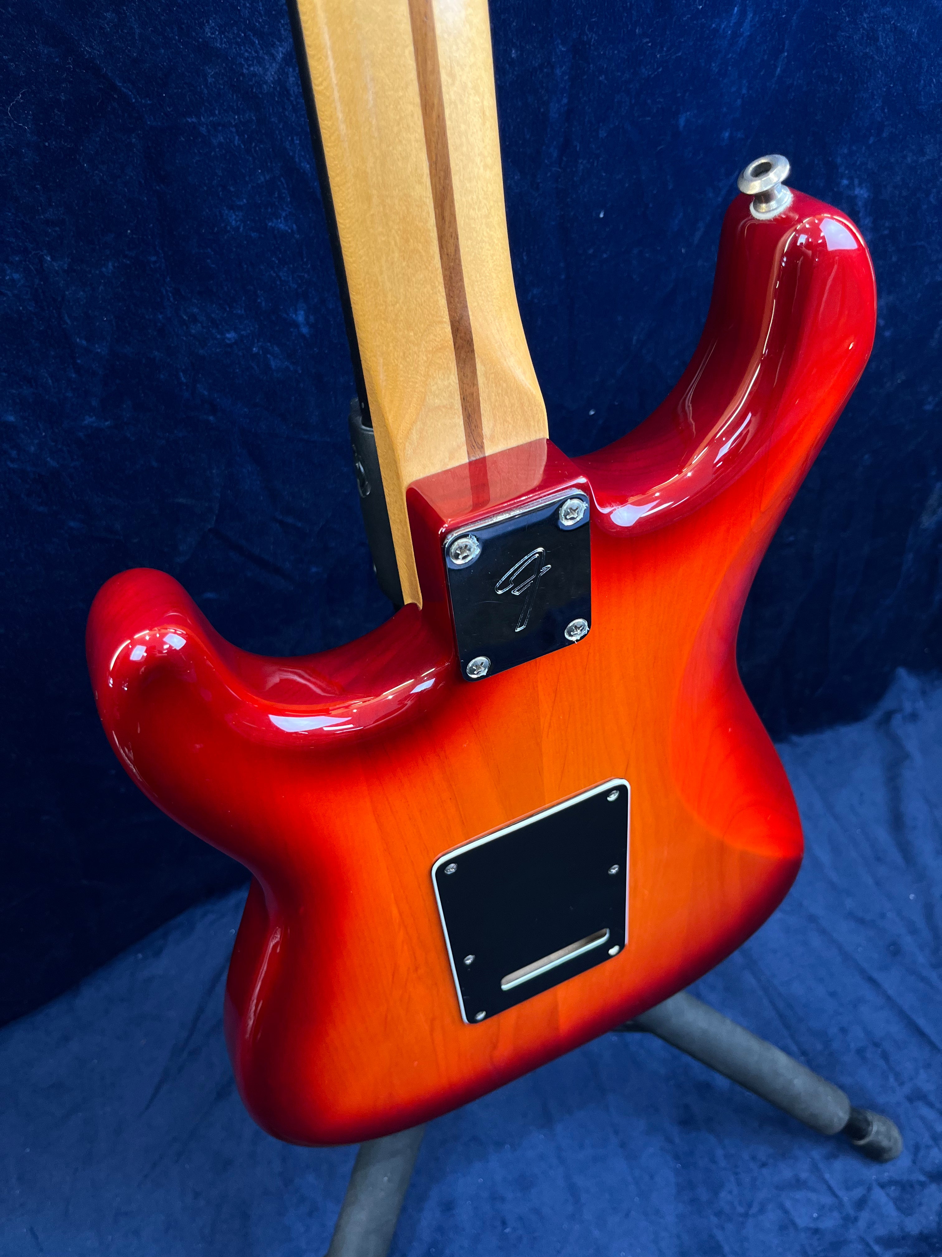 Guitare électrique avec un corps rouge dégradé, manche bois avec bande sombre, plaque noire au dos, posée sur un support contre fond bleu.