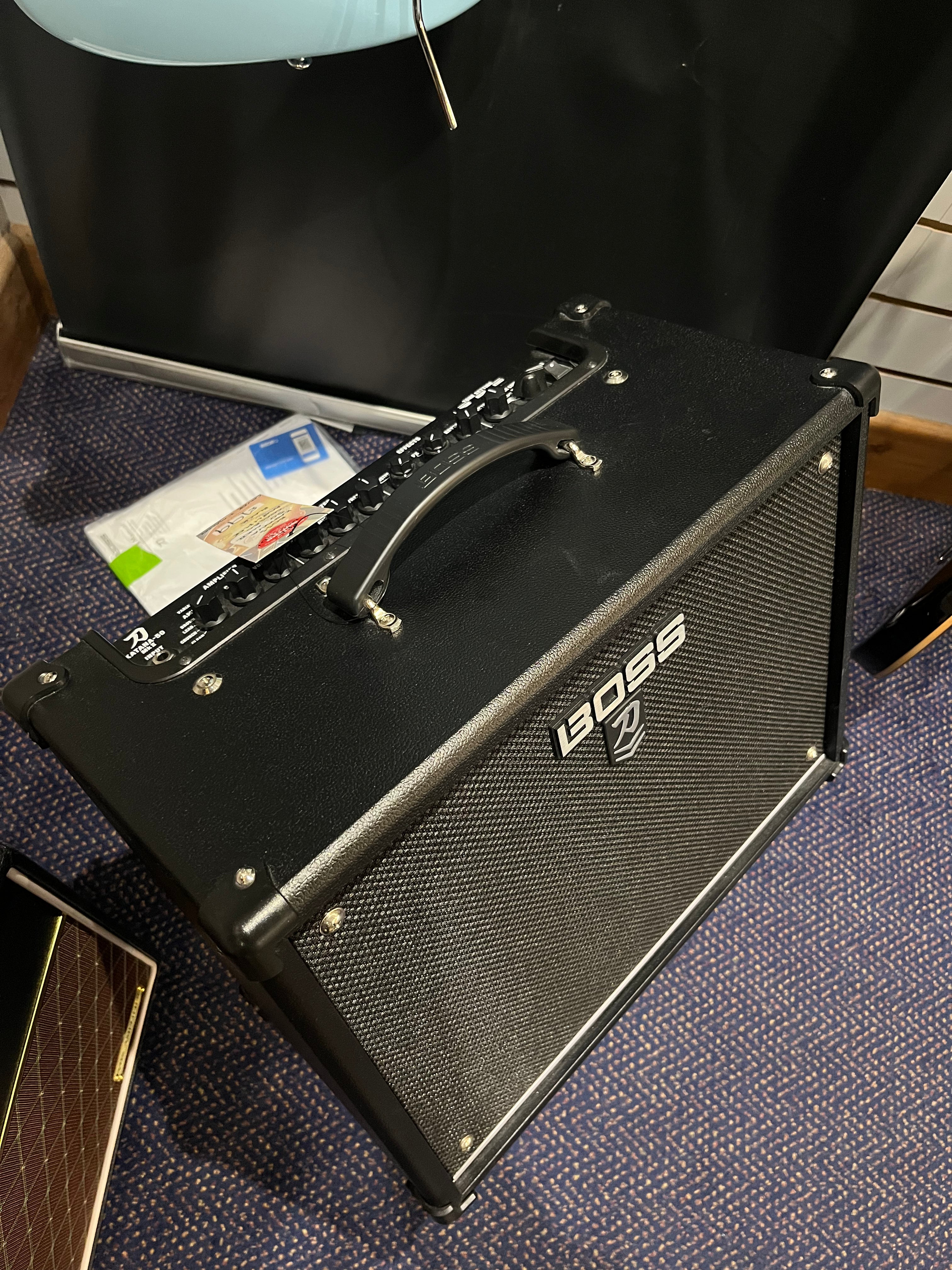 Black Boss Katana guitar amplifier with control knobs and handle on top, angled on a carpeted floor near a hanging guitar.