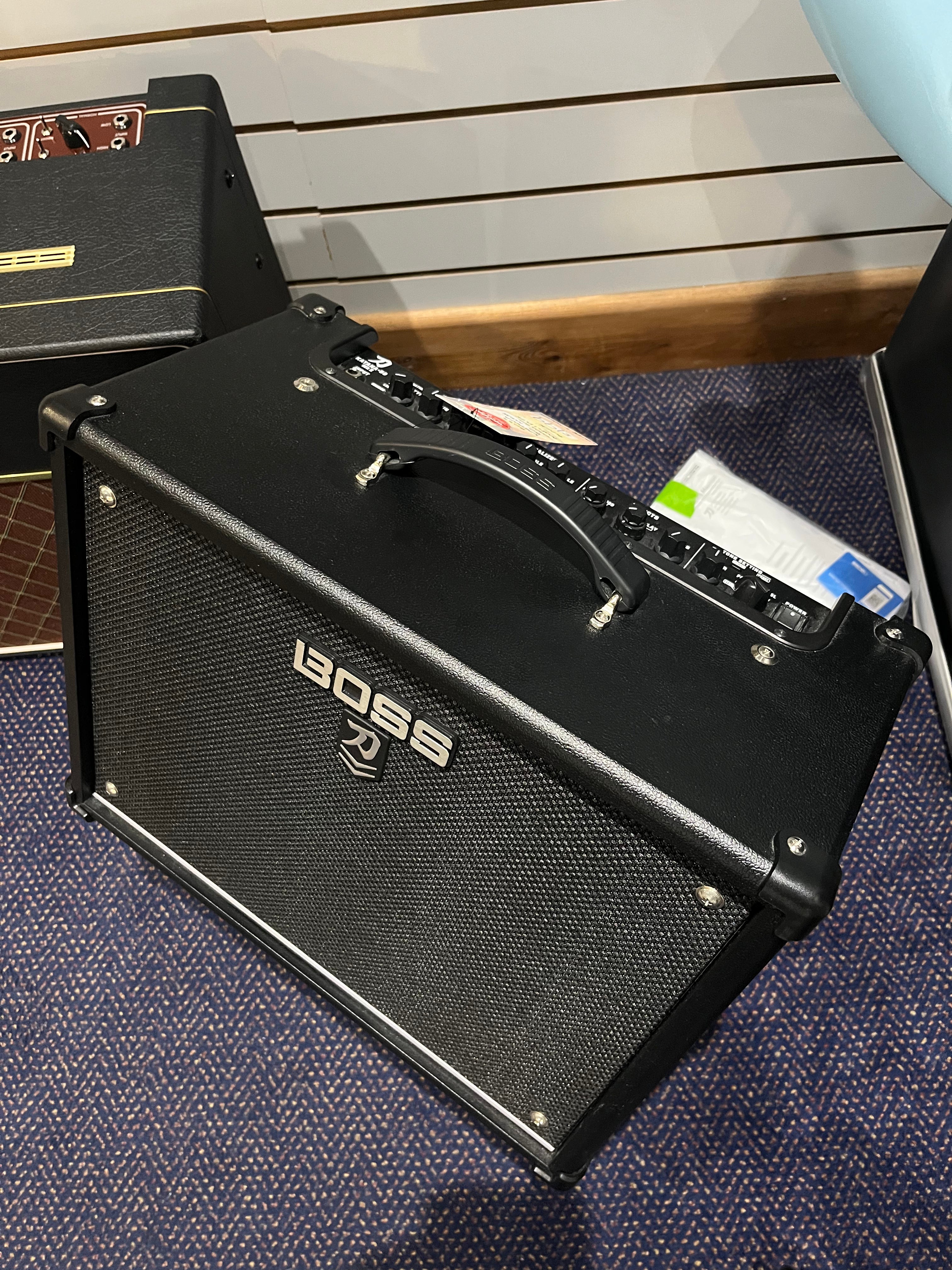 Black Boss guitar amplifier with a top carry handle, control knobs on top, and a textured front grille, sitting on a carpeted floor.