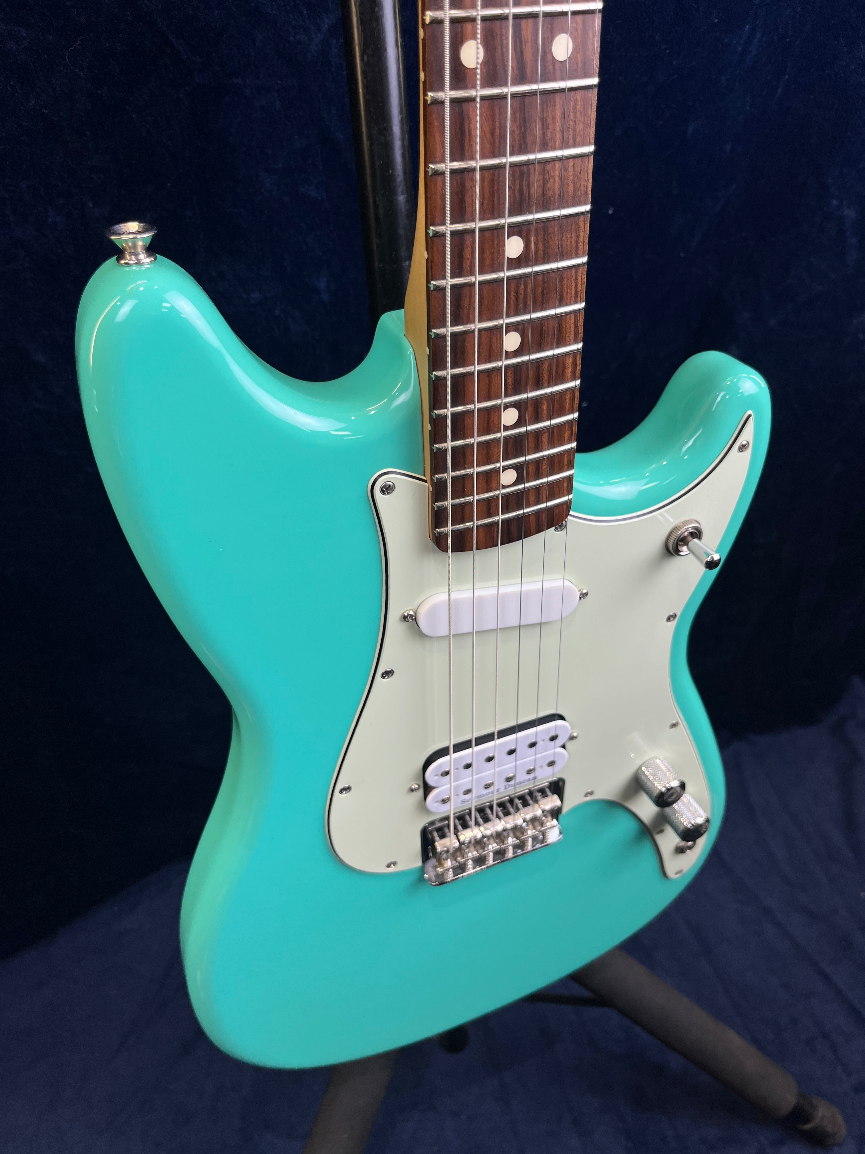 Turquoise electric guitar with white pickguard, two pickups, one knob, and a pickup switch, shown on a stand against a dark background.