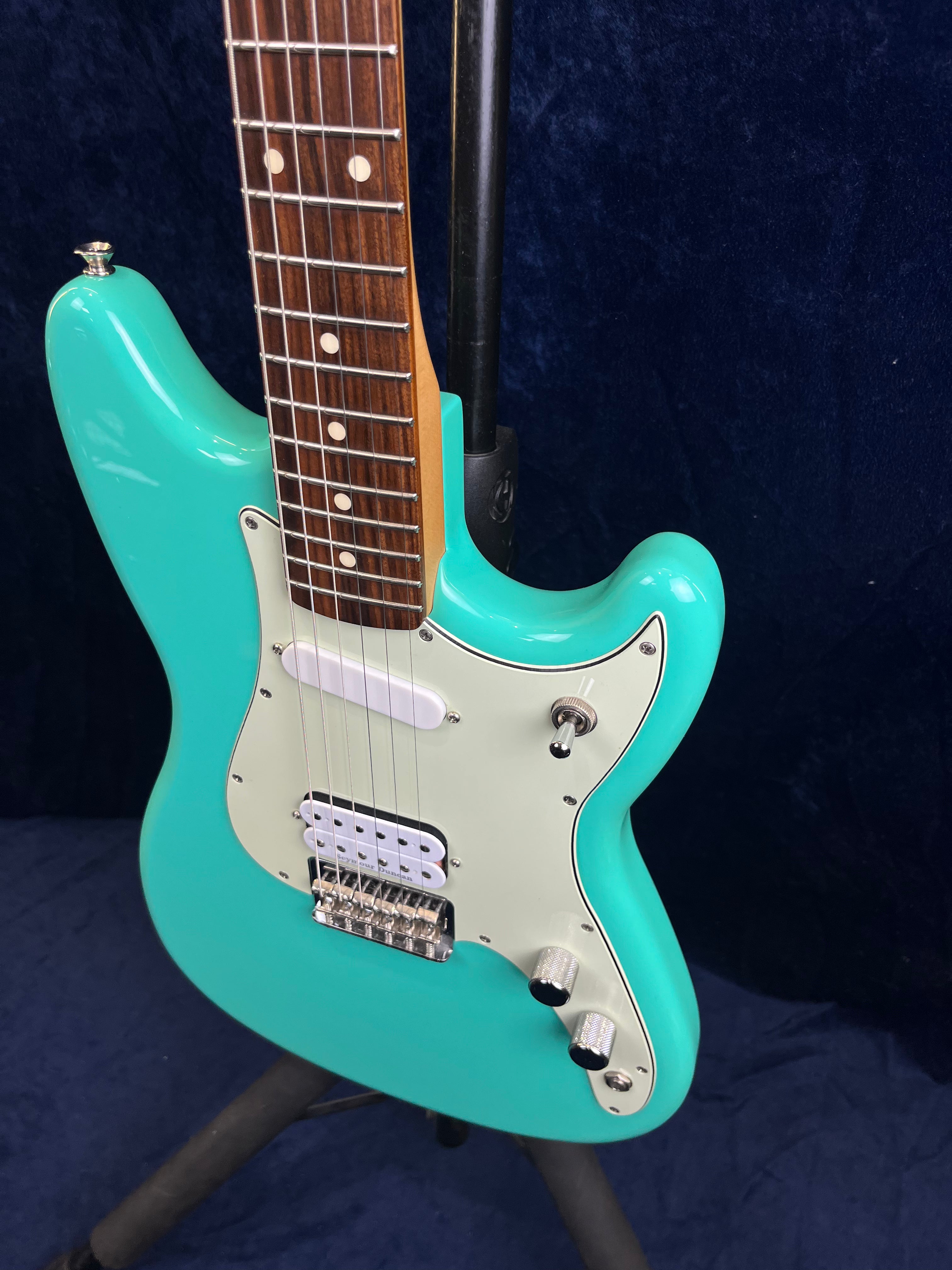 Turquoise electric guitar with a white pickguard, chrome knobs, and a rosewood fretboard standing on a black guitar stand.
