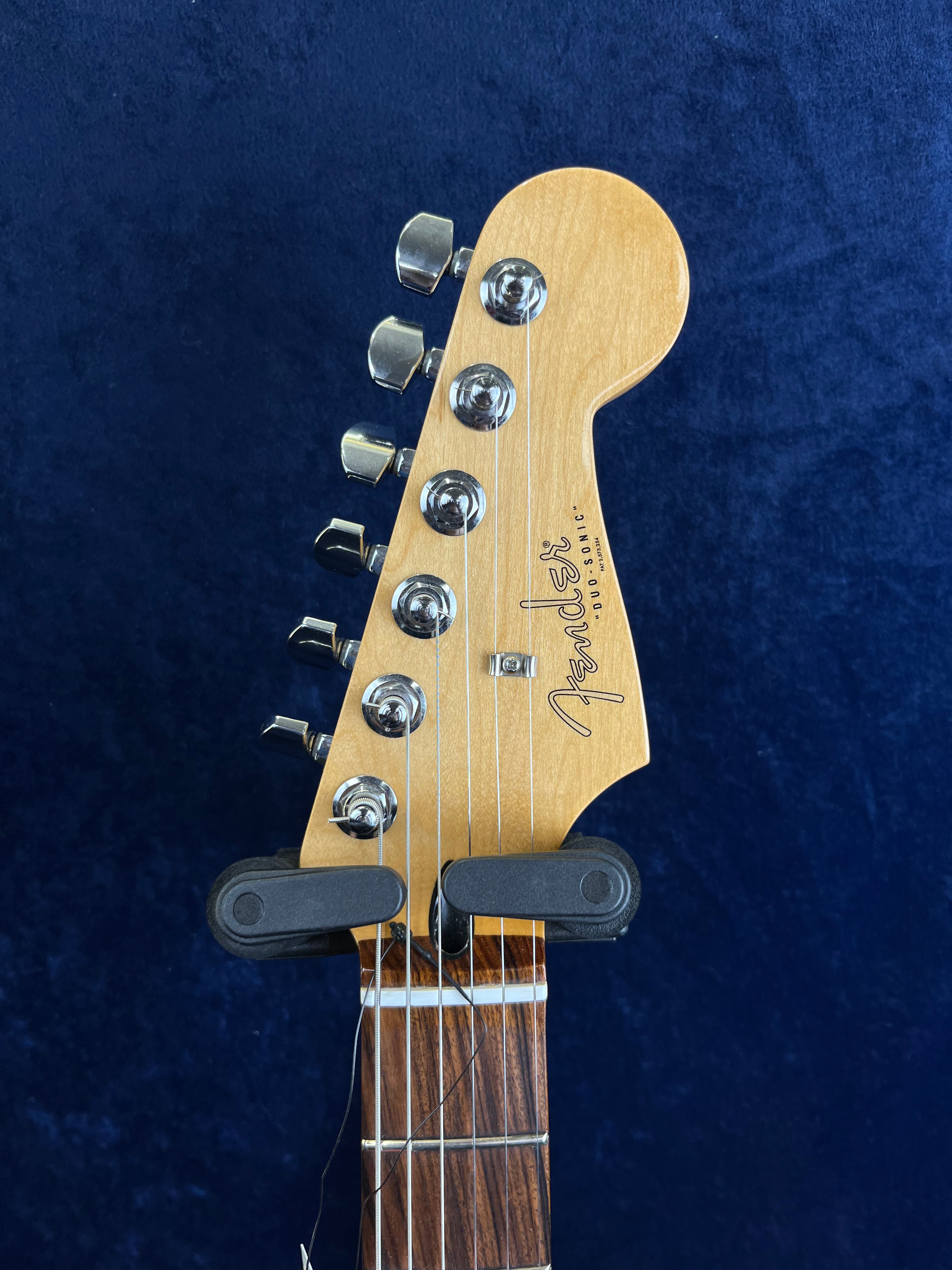 Close-up of a Fender Duo-Sonic electric guitar headstock with six tuning pegs, visible strings, and part of the neck against a dark blue background.