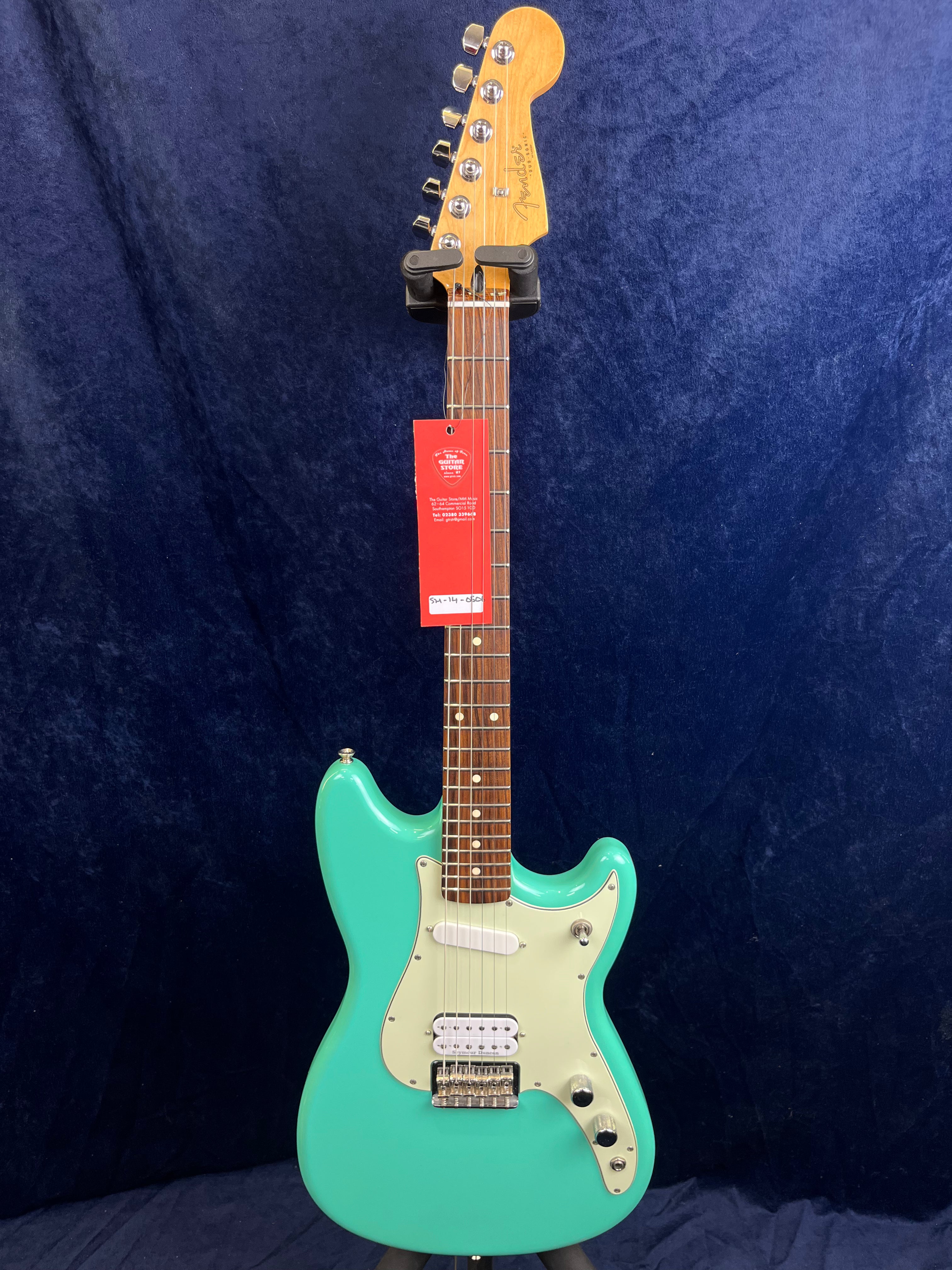 A seafoam green electric guitar with a white pickguard, two pickups, a wooden fretboard, and chrome tuning pegs hangs against a dark blue background.