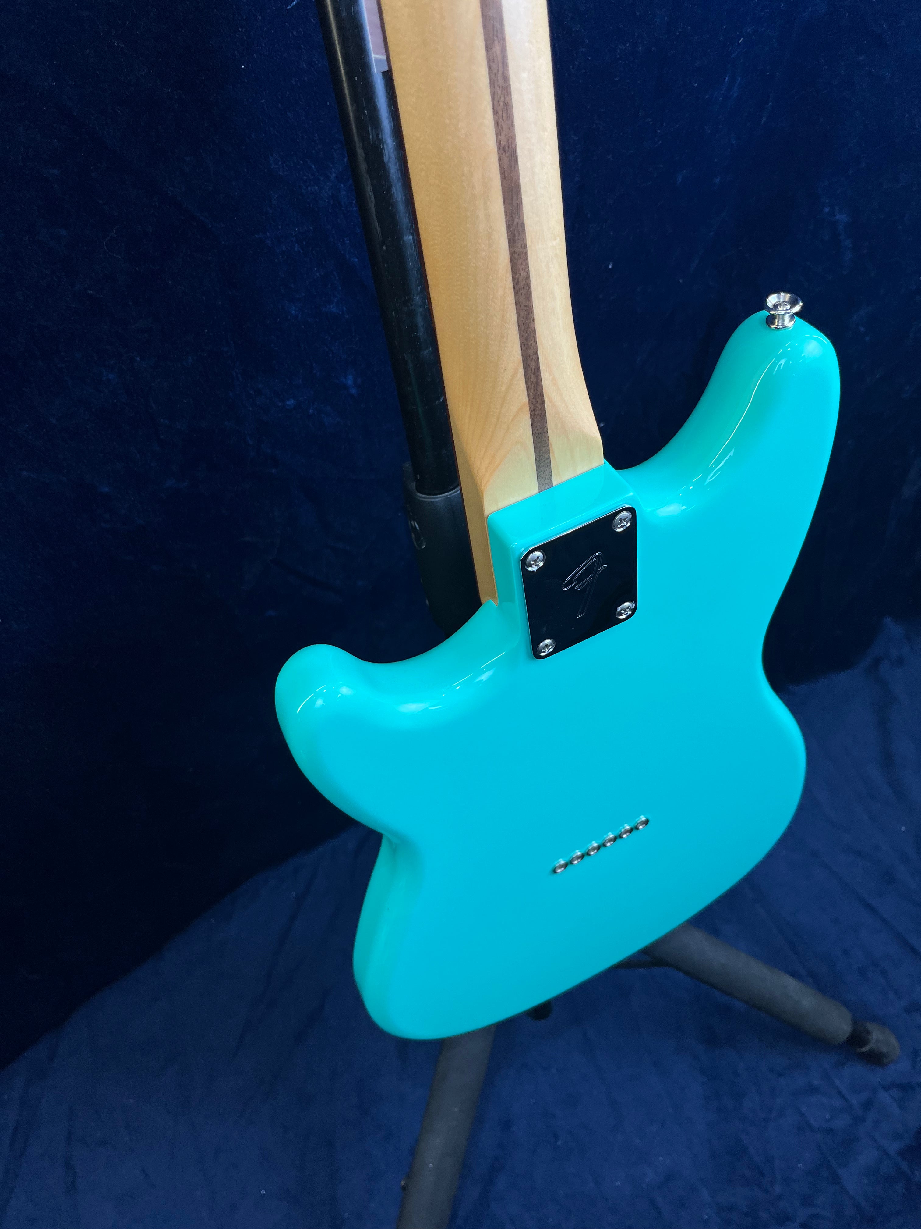 A turquoise electric guitar rests on a stand, showing its smooth back, bolt-on neck with a dark stripe, and six string ferrules aligned in a row.