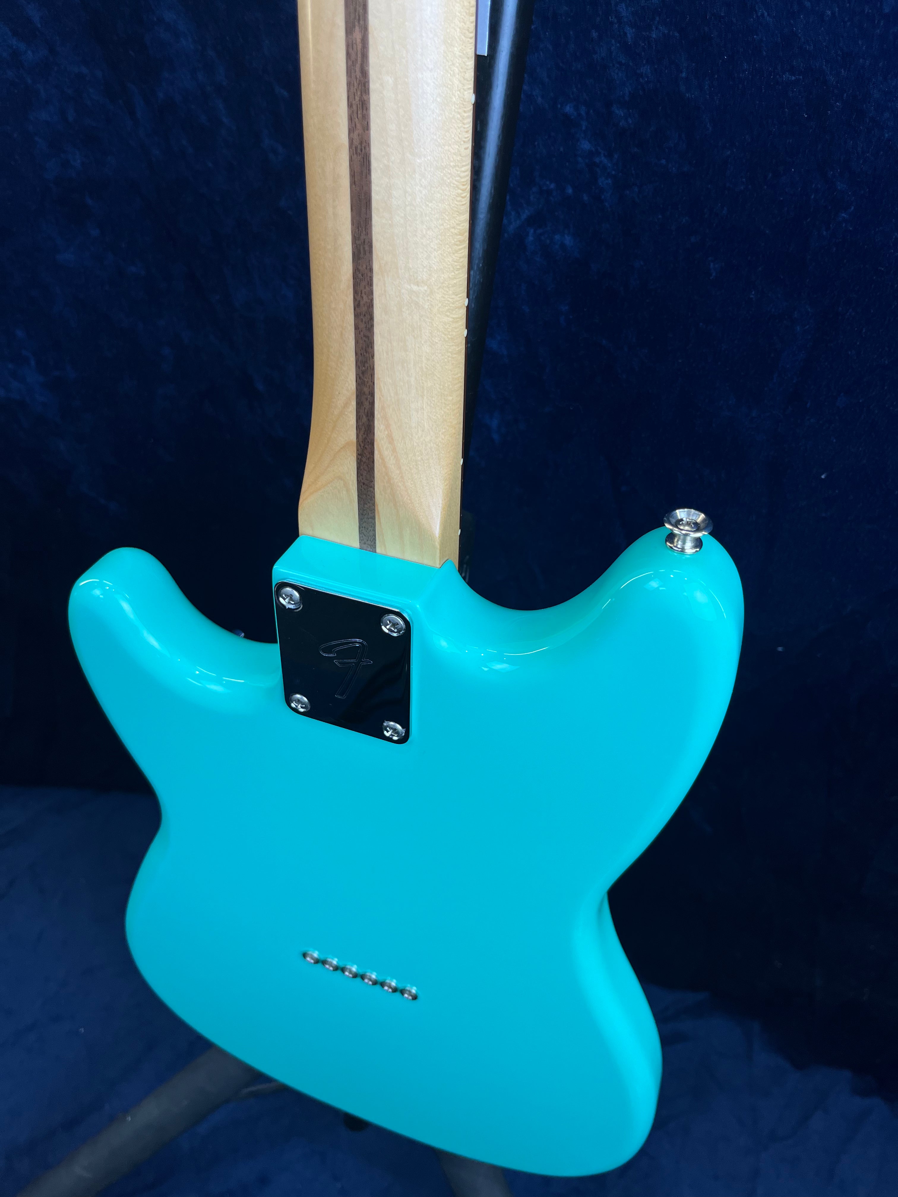 A turquoise electric guitar shown from the back with a natural wood neck, a dark stripe down the neck, and a chrome neck plate with an engraved "F".