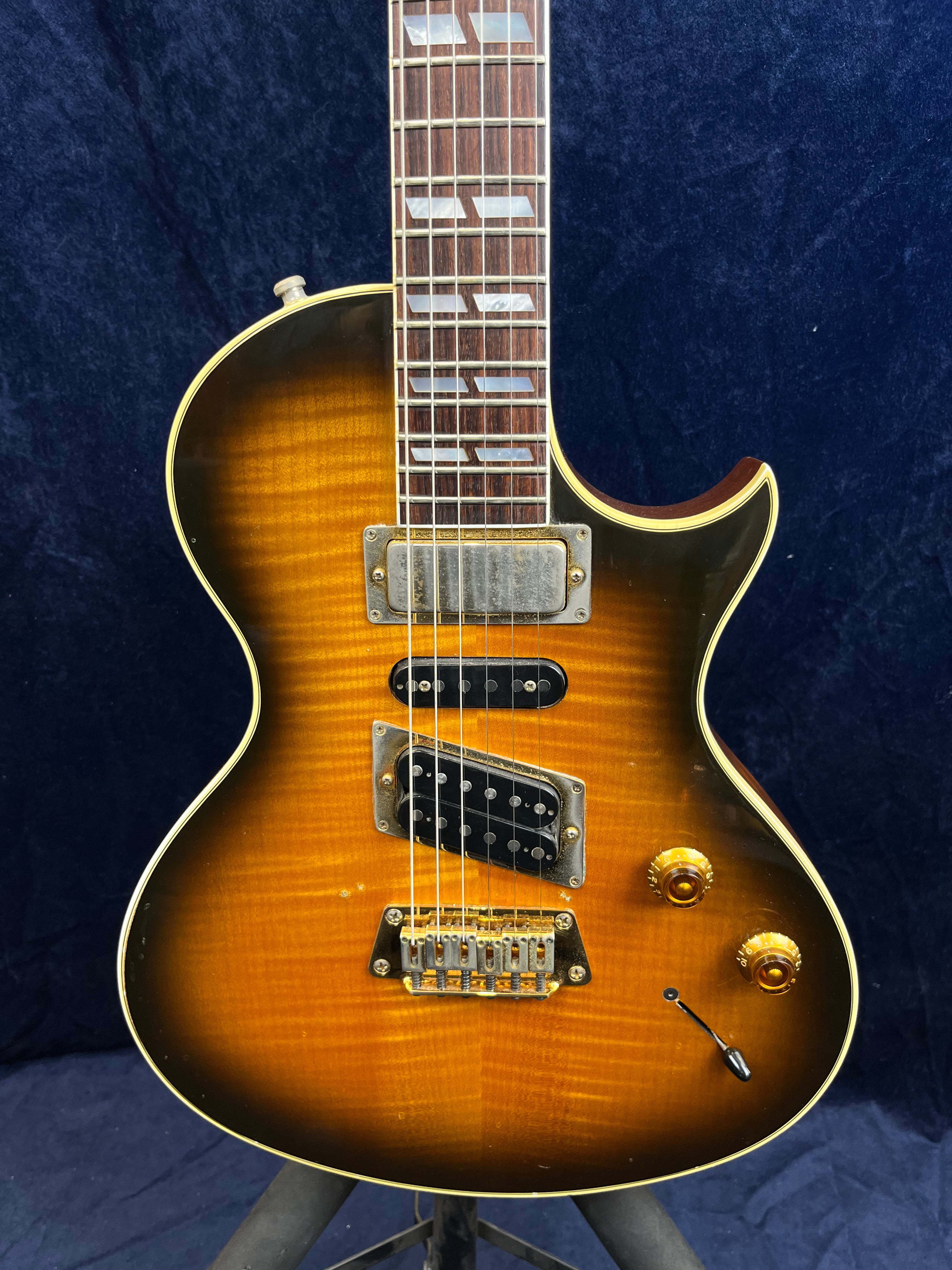 Sunburst Gibson electric guitar with three pickups, two gold control knobs, and a switch on the lower right, featuring a single cutaway body.