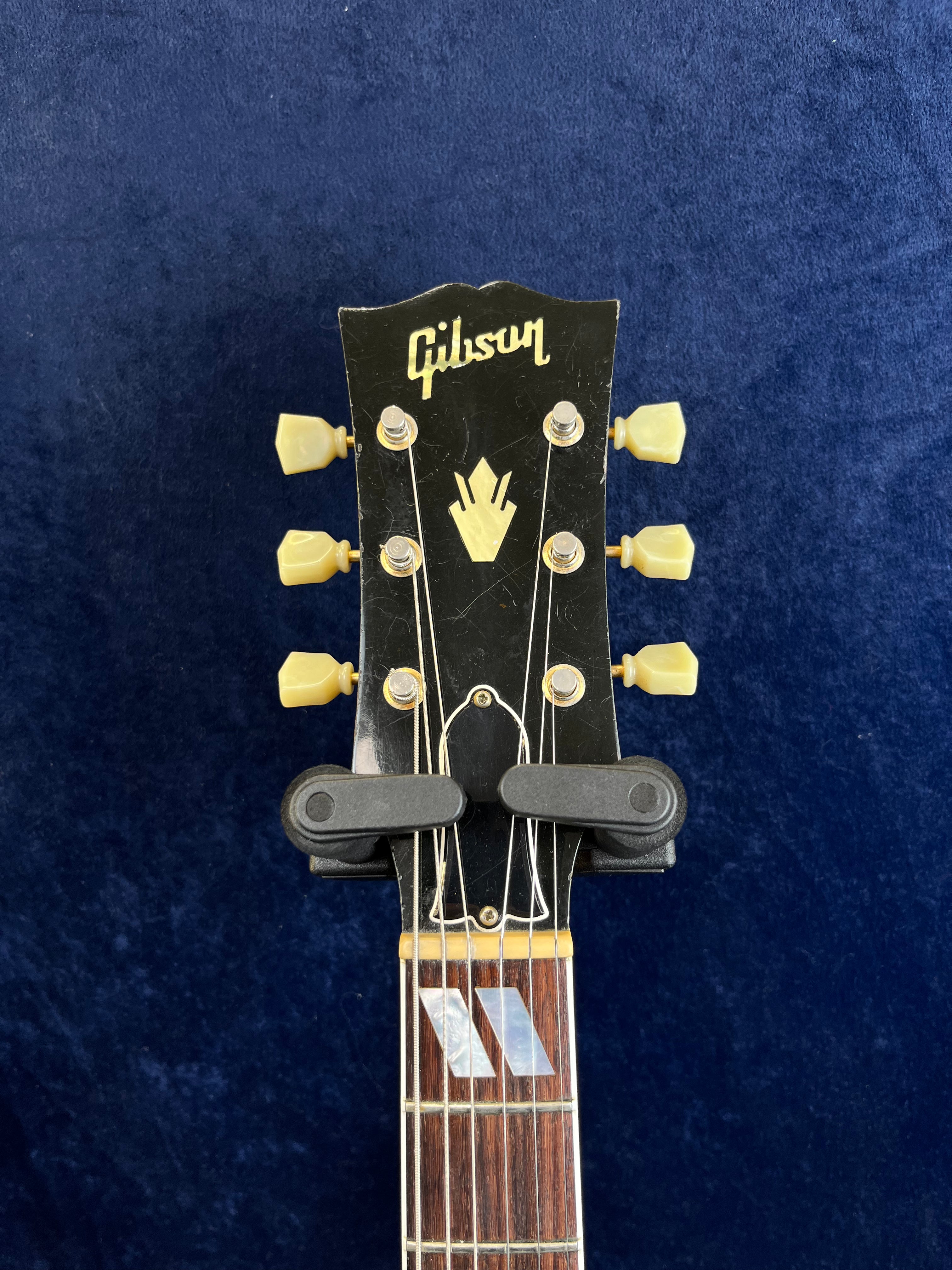 Gibson guitar headstock with six cream tuning pegs, gold logo, and decorative inlay, against a dark blue background.