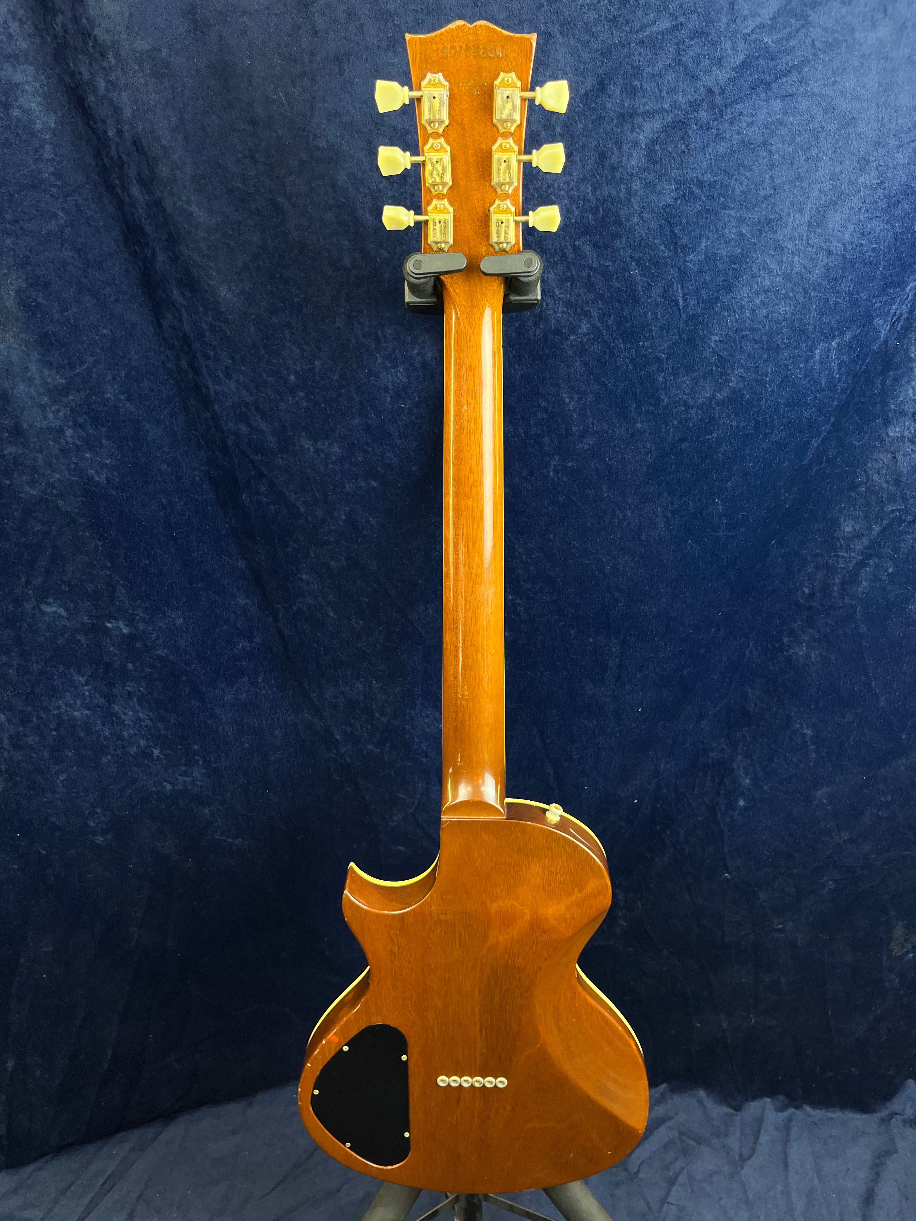 The back of a brown Gibson electric guitar, showing six tuning pegs, a black electronics cover, and wood grain against a dark blue background.