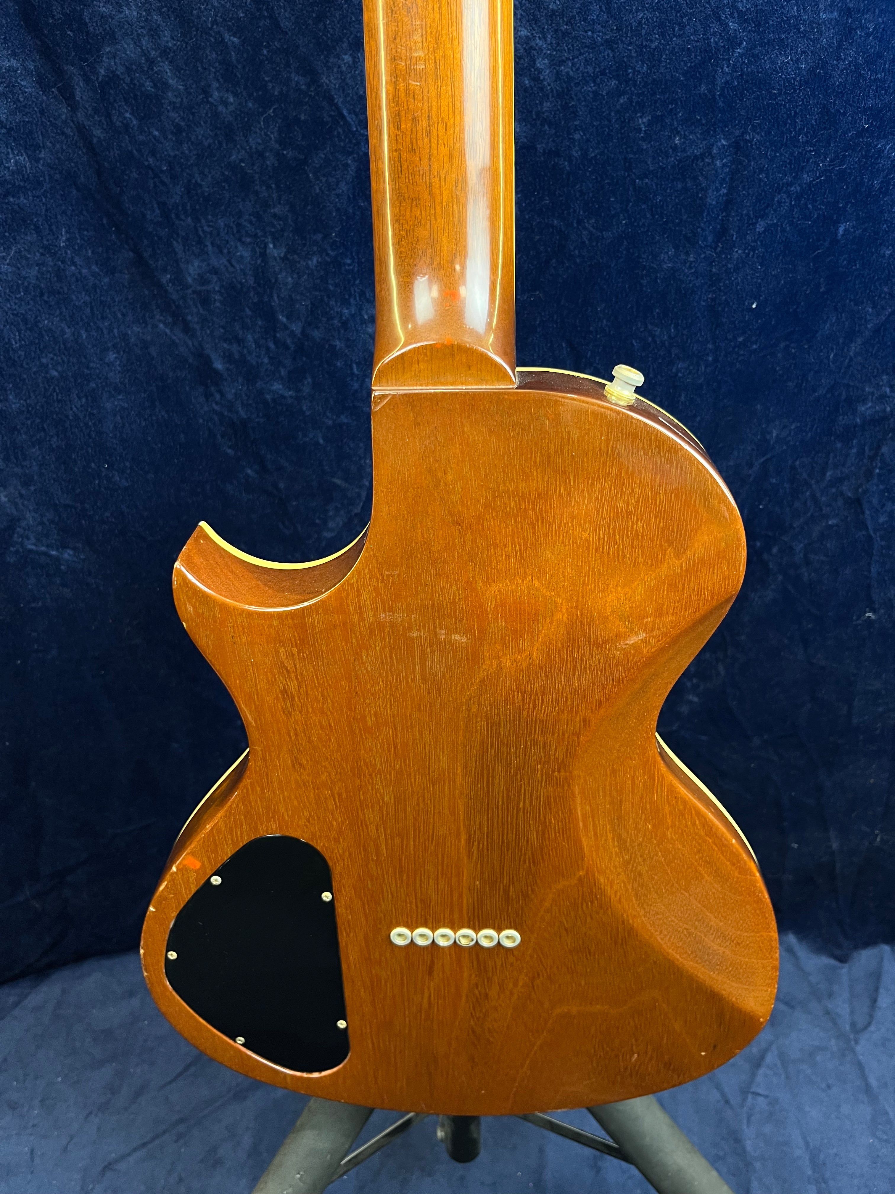 Rear view of a brown Gibson electric guitar body showing wood grain, black electronics cover, six string ferrules, and a strap button.
