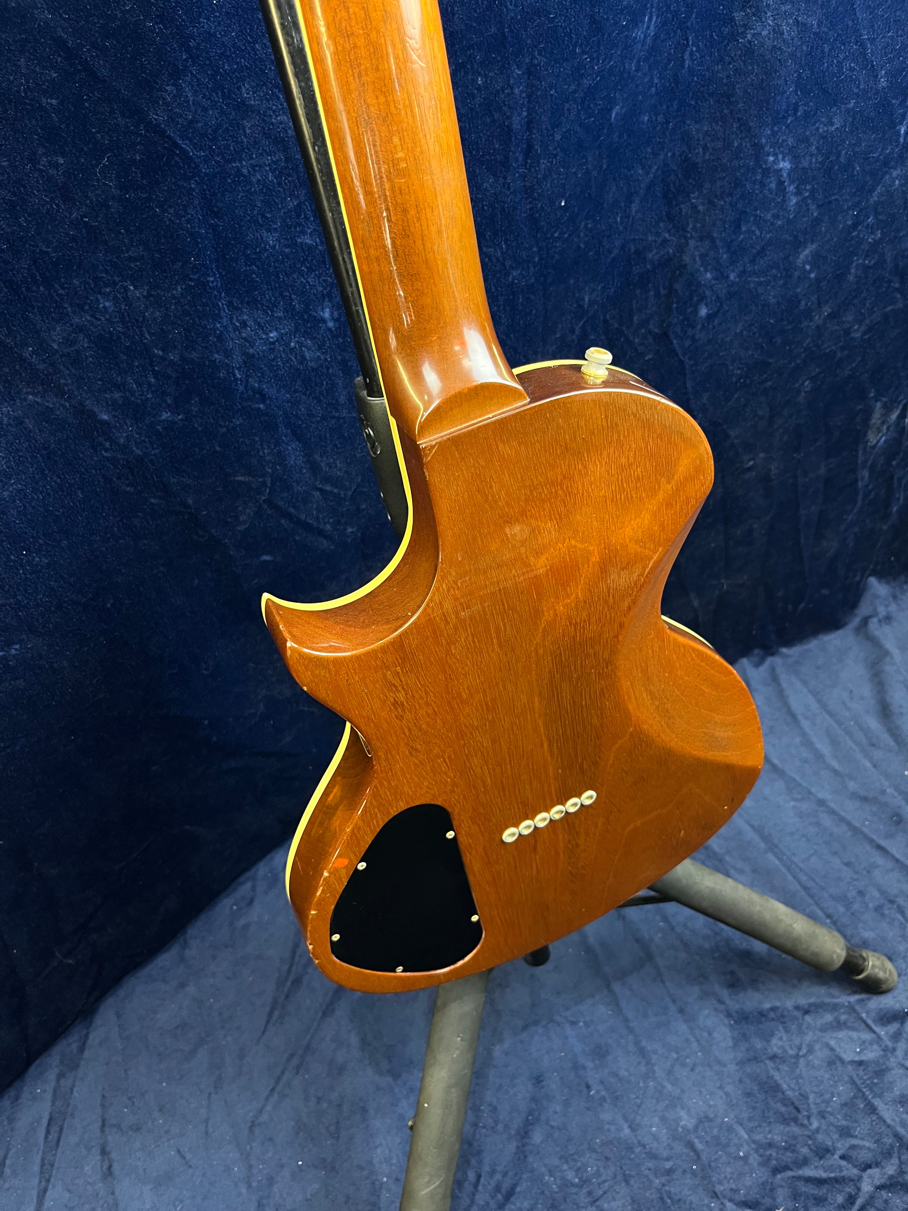 Back view of a single-cutaway Gibson electric guitar with a brown wood finish, black electronics cover, and cream binding along the body and neck.