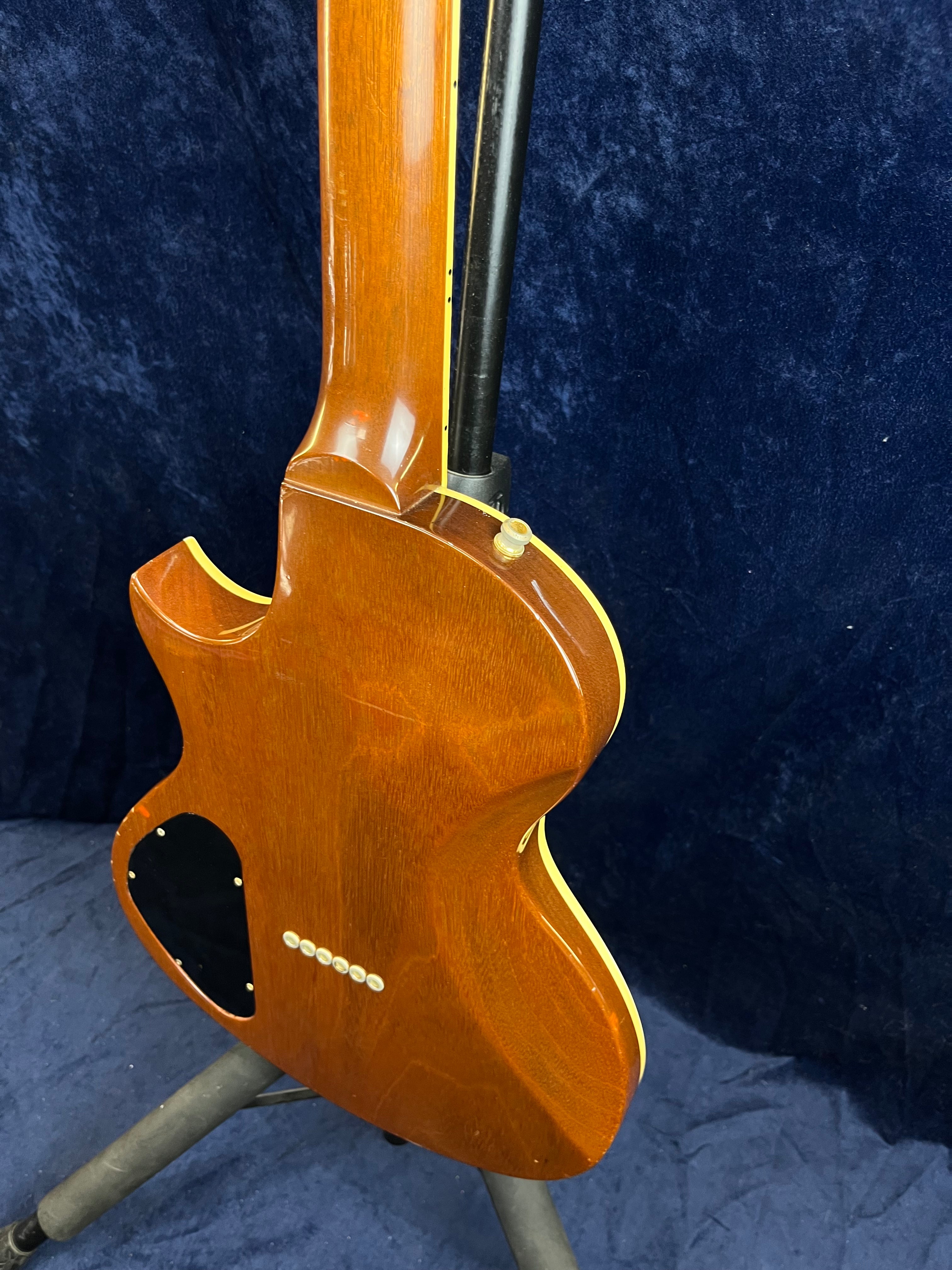 Rear view of a Gibson electric guitar with a brown wood finish, cream binding, a black electronics cover, and six string ferrules on the body.
