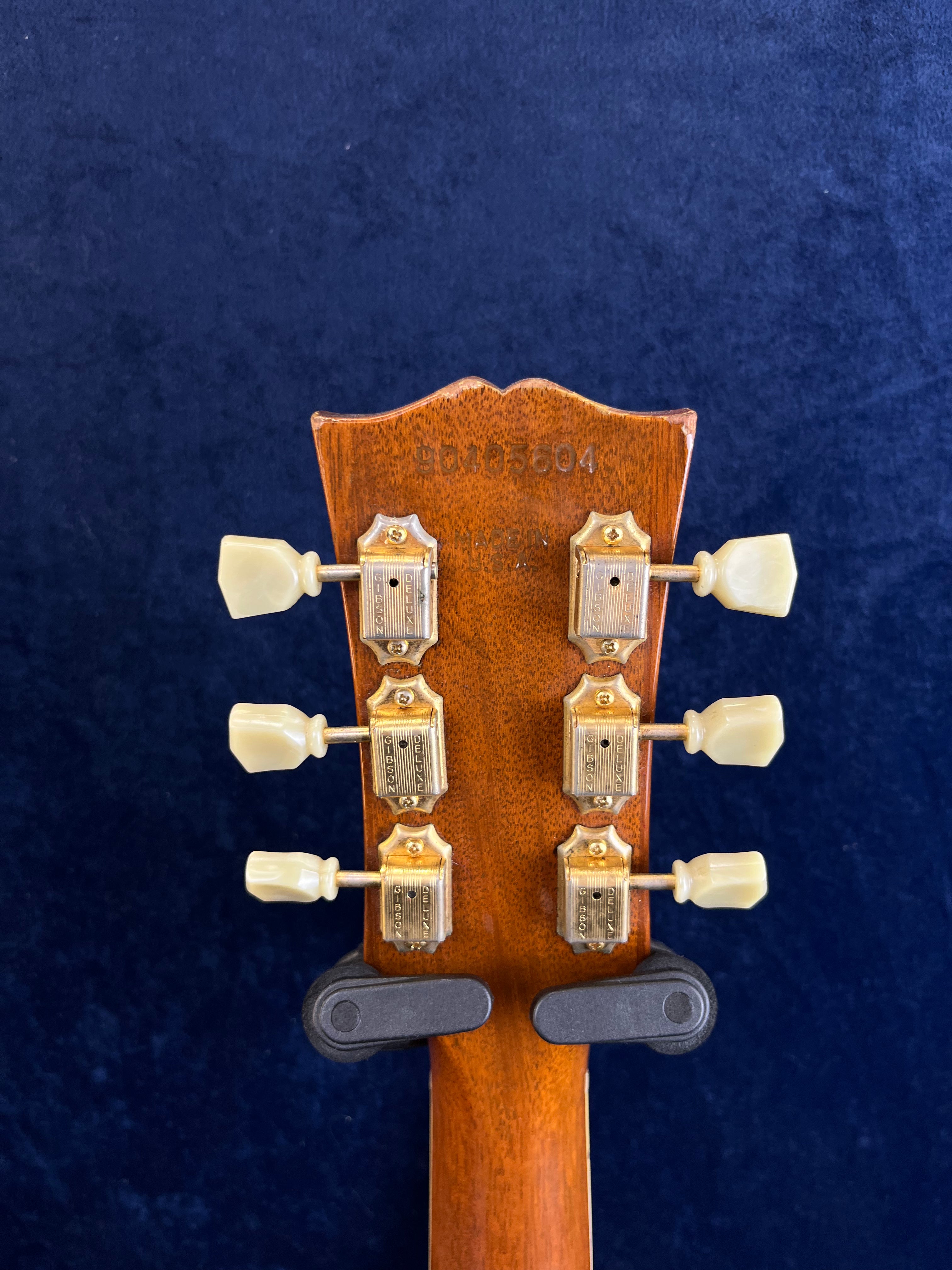 Back of a Gibson guitar headstock with six vintage-style tuning pegs, serial number stamped at the top on brown wood, held upright in a hanger.