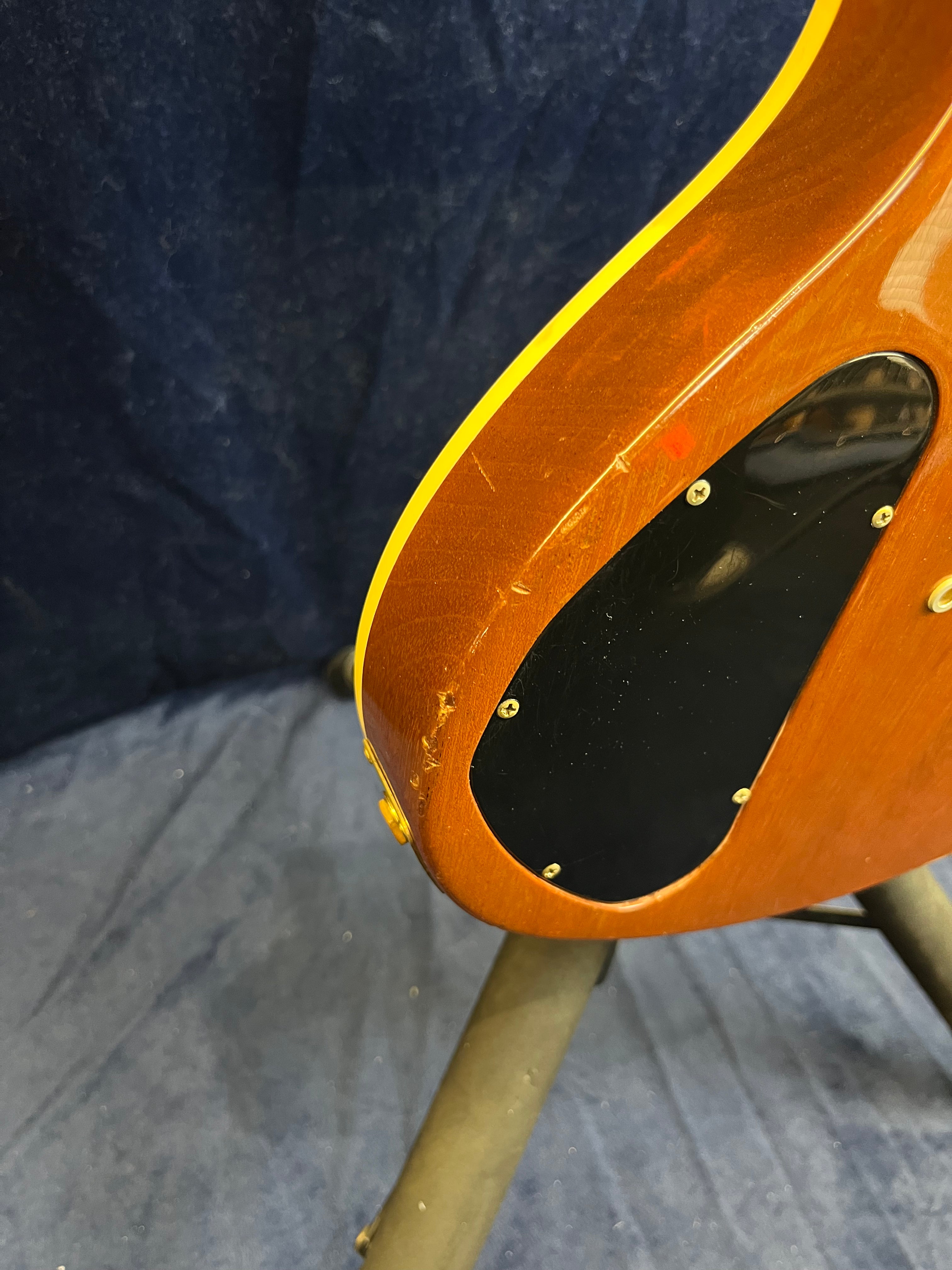 Back corner of a Gibson guitar with a brown wood finish, yellow trim, black plastic electronics cover, and visible scratches and dings on the edge.