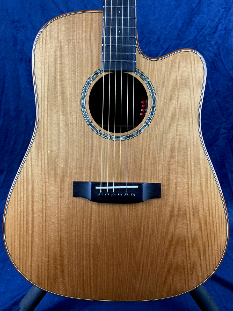 Natural wood acoustic guitar with cutaway body, six strings, round sound hole with decorative inlay, and black bridge against a dark blue background.
