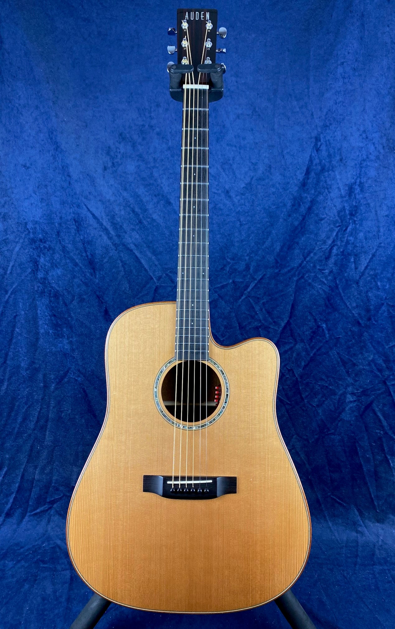 Auden acoustic guitar with a natural wood finish, cutaway body shape, six strings, and black tuning pegs, displayed against a blue background.