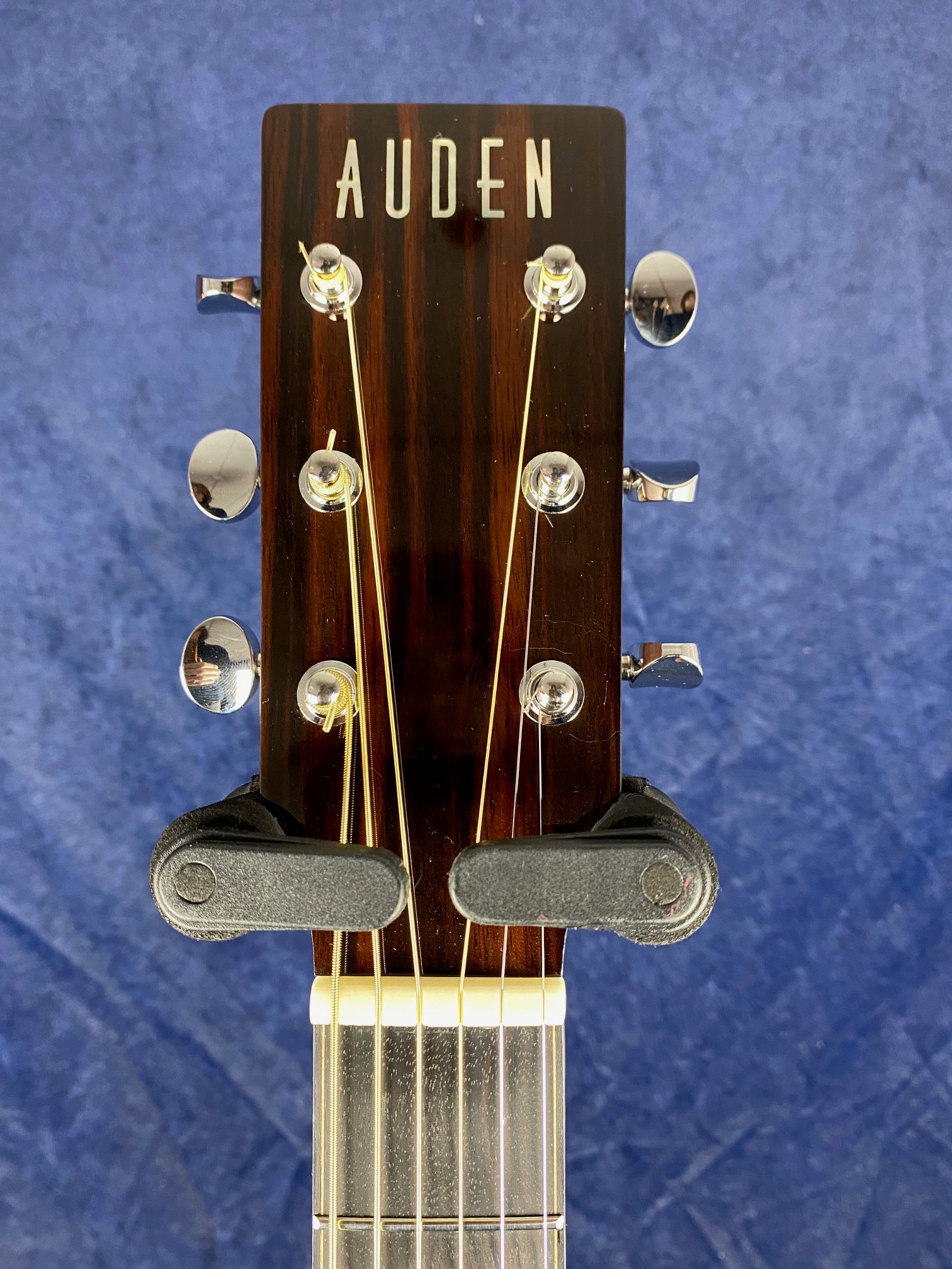 Auden acoustic guitar headstock with six tuning pegs and gold strings, held by a black hanger against a blue background.