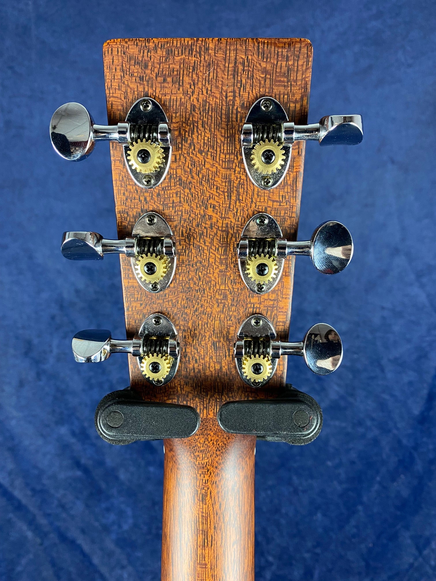 Six chrome tuning pegs with exposed gears on a wooden acoustic guitar headstock, shown from the back against a blue background.