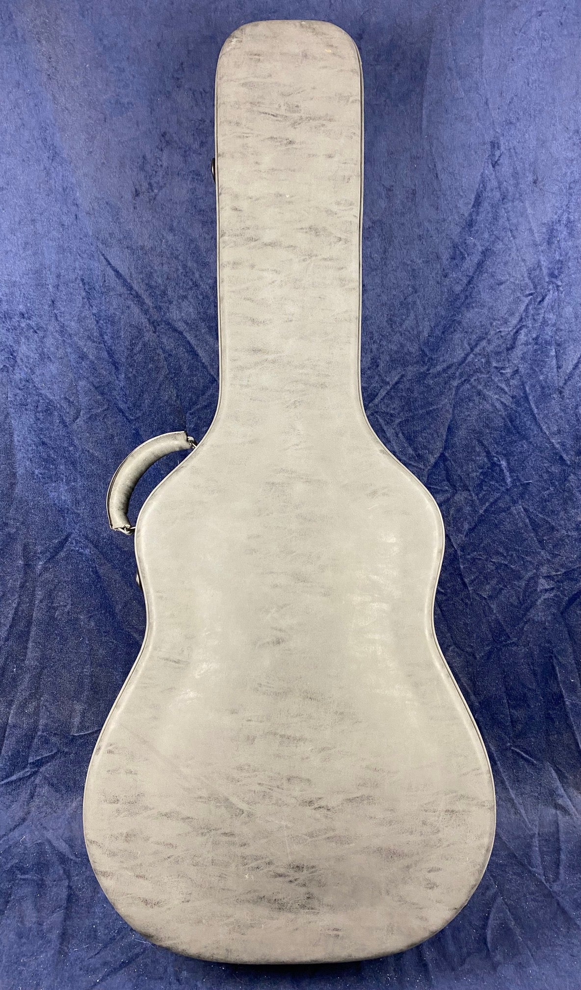 Closed, light gray acoustic guitar case with a curved handle on the left side, lying on a textured blue background.