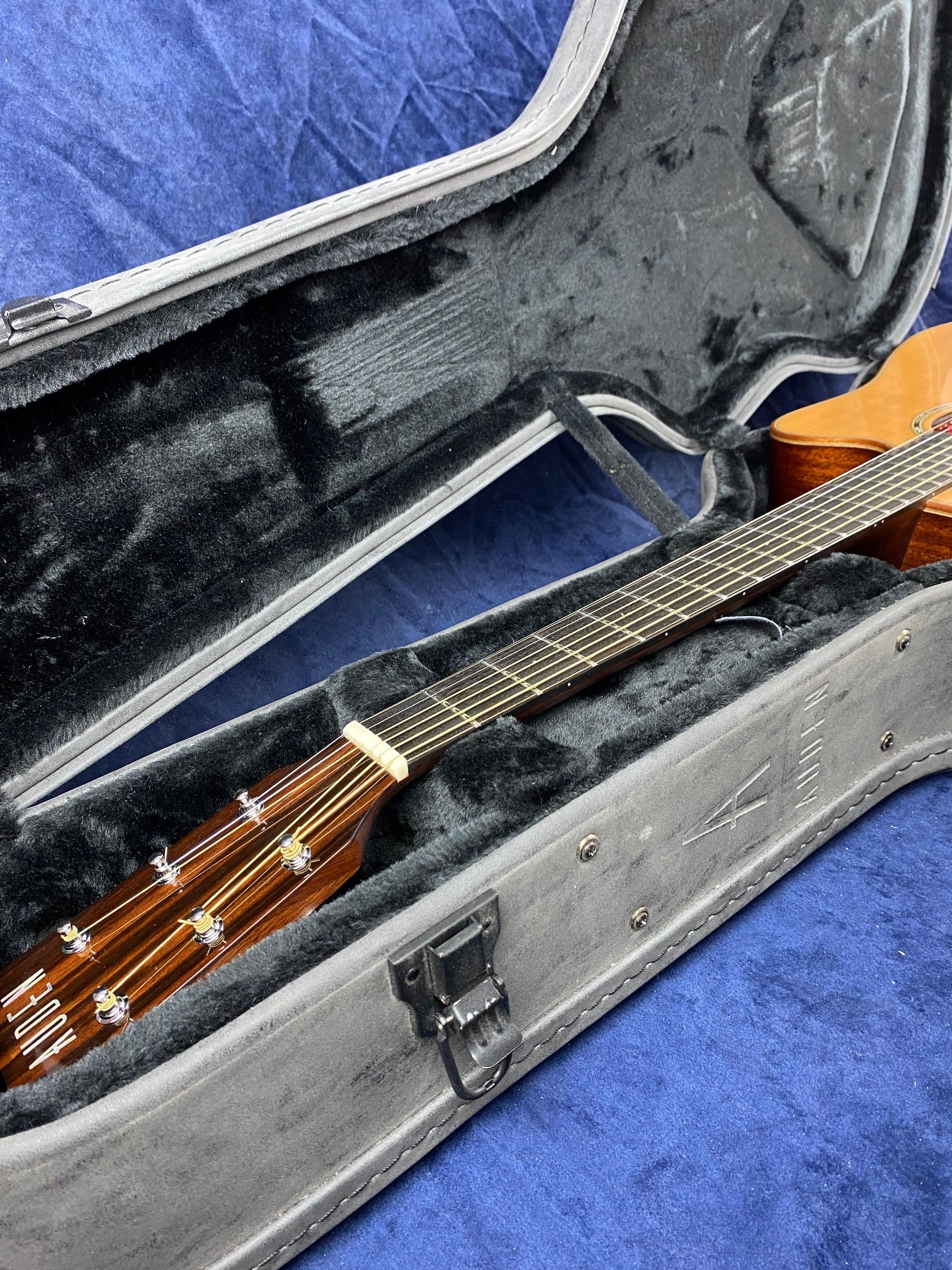 Wooden acoustic guitar rests inside a black plush-lined hard case, shown partially open on a textured blue surface.