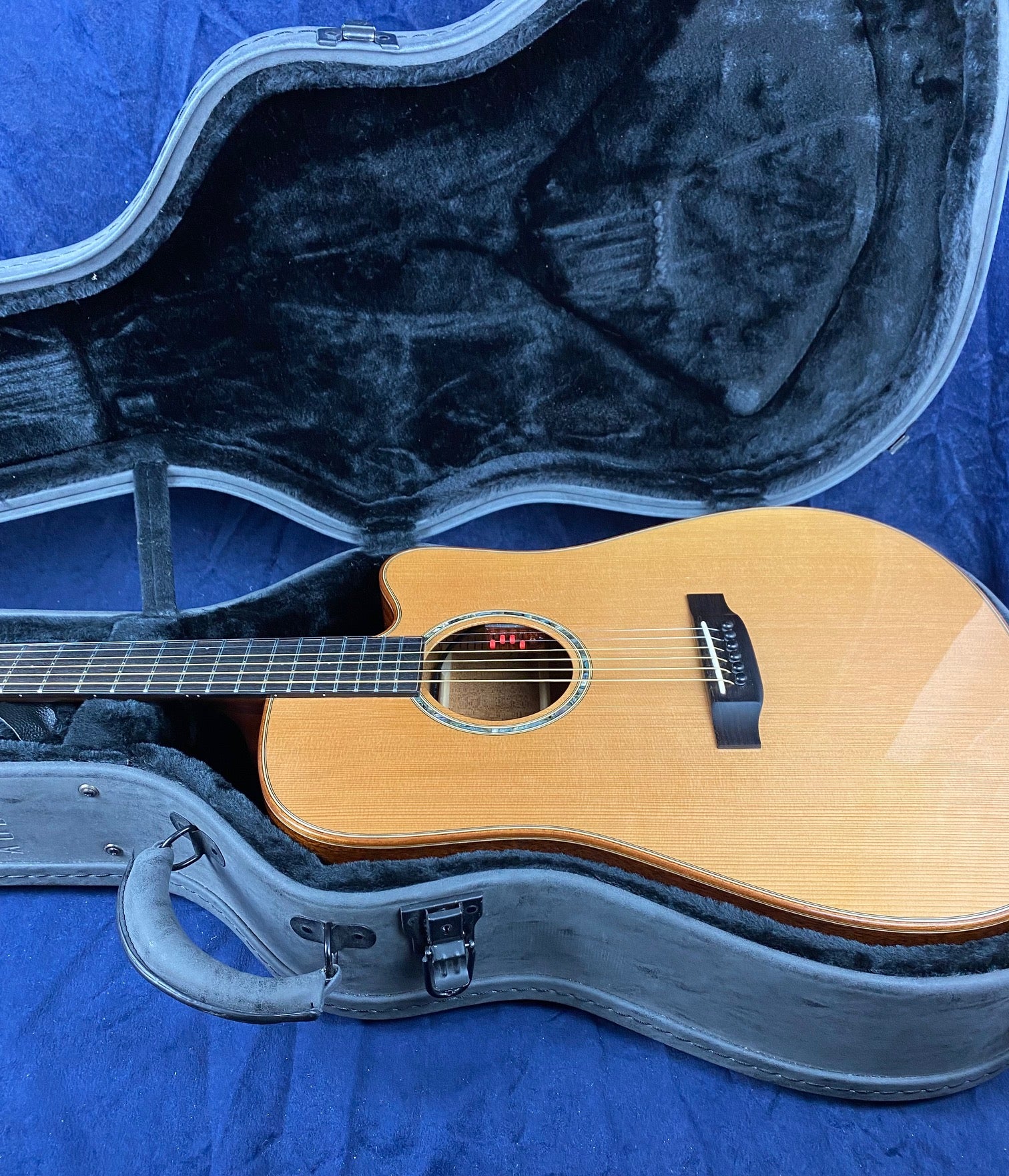 Wooden acoustic guitar with a cutaway body resting inside a black, plush-lined hard case on a blue surface.