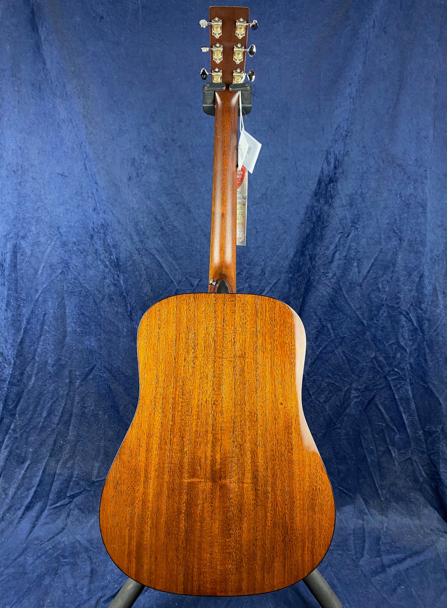Brown acoustic guitar shown from the back, displaying its wooden grain, straight neck, and silver tuning pegs against a dark blue background.