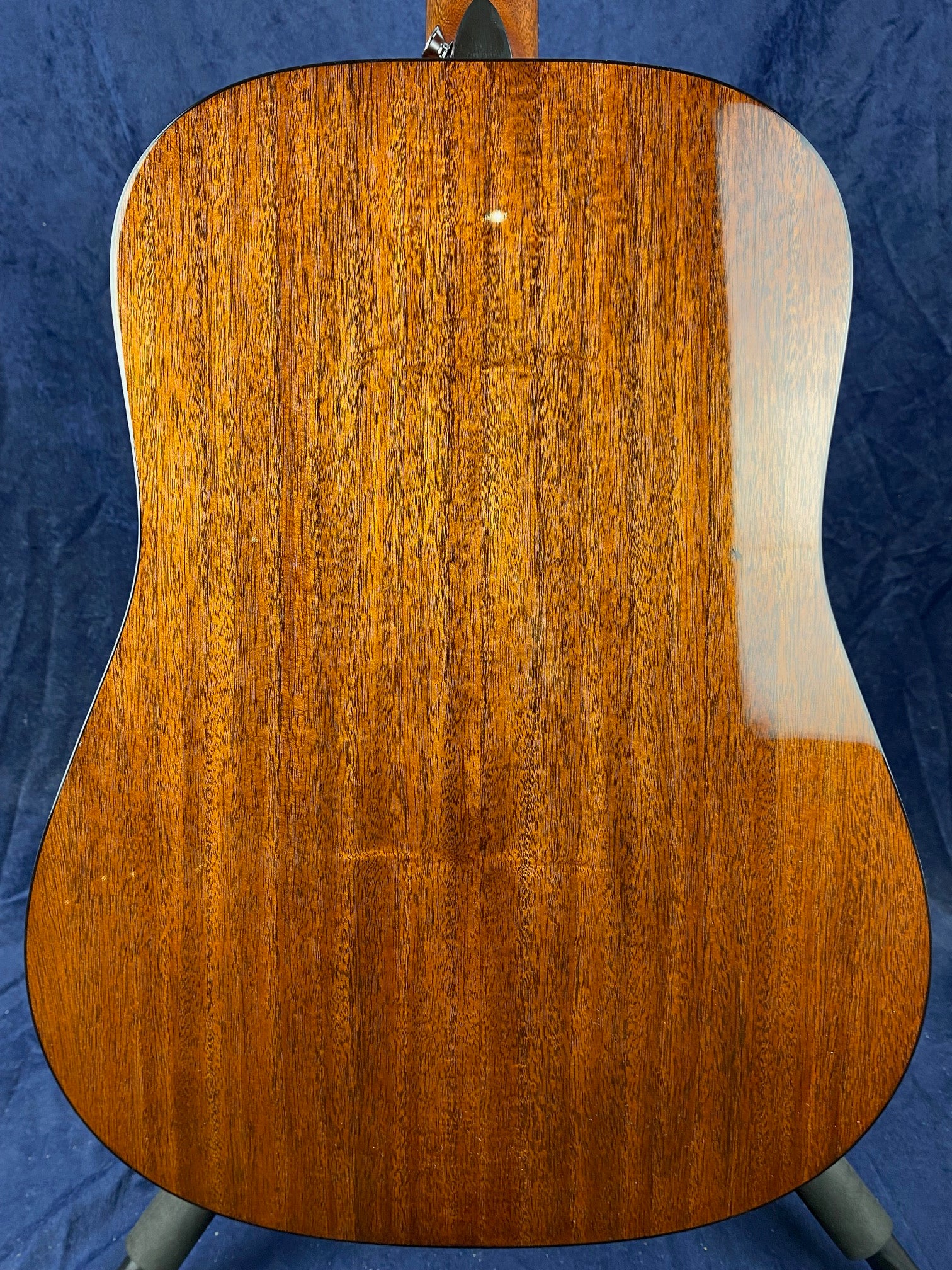 Back of an acoustic guitar showing polished, dark brown wood with a vertical grain pattern and rounded edges, set against a deep blue background.