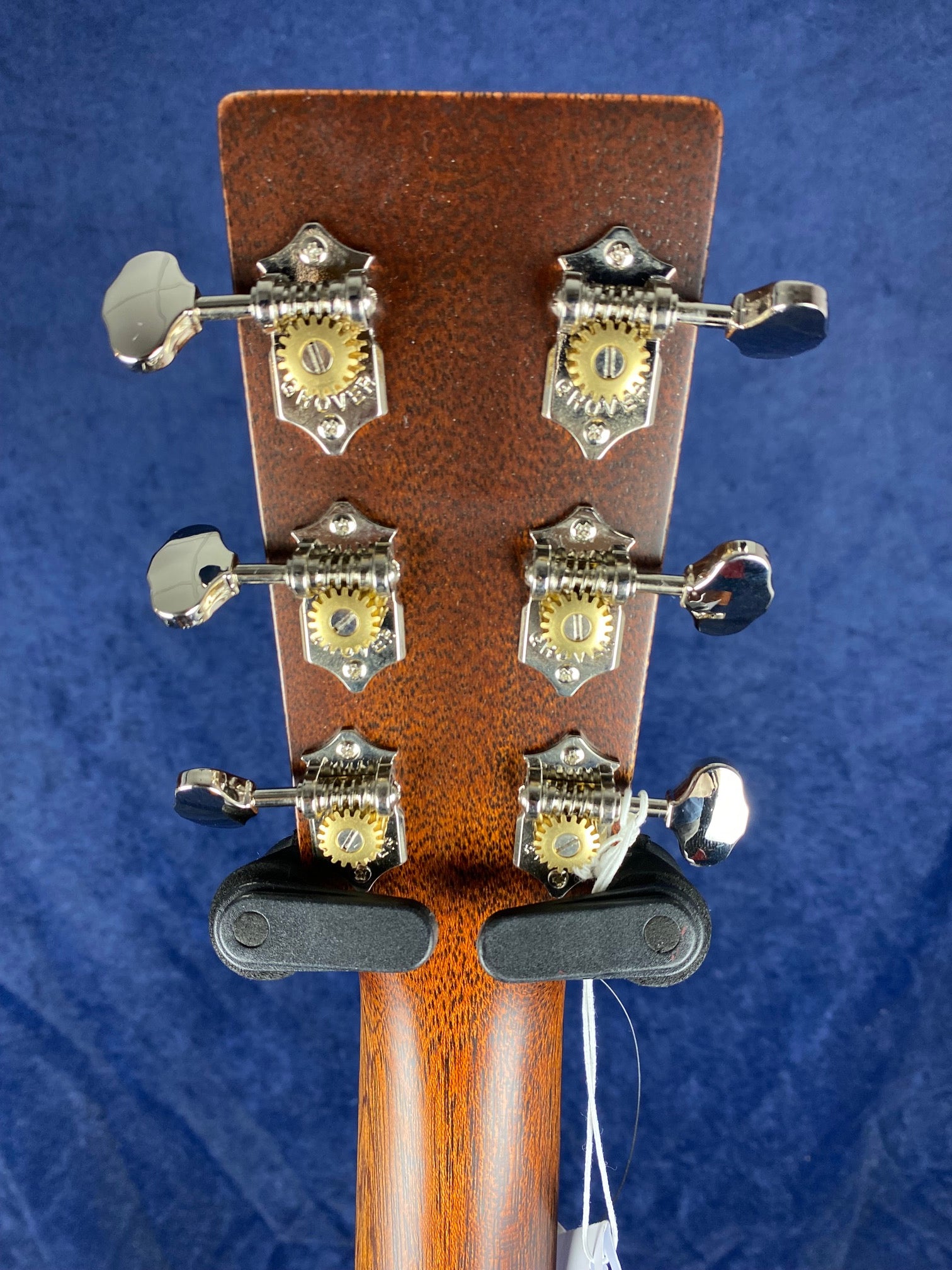 Six chrome tuning pegs with decorative gear plates are mounted on the back of a wooden acoustic guitar headstock, positioned against a dark blue background.