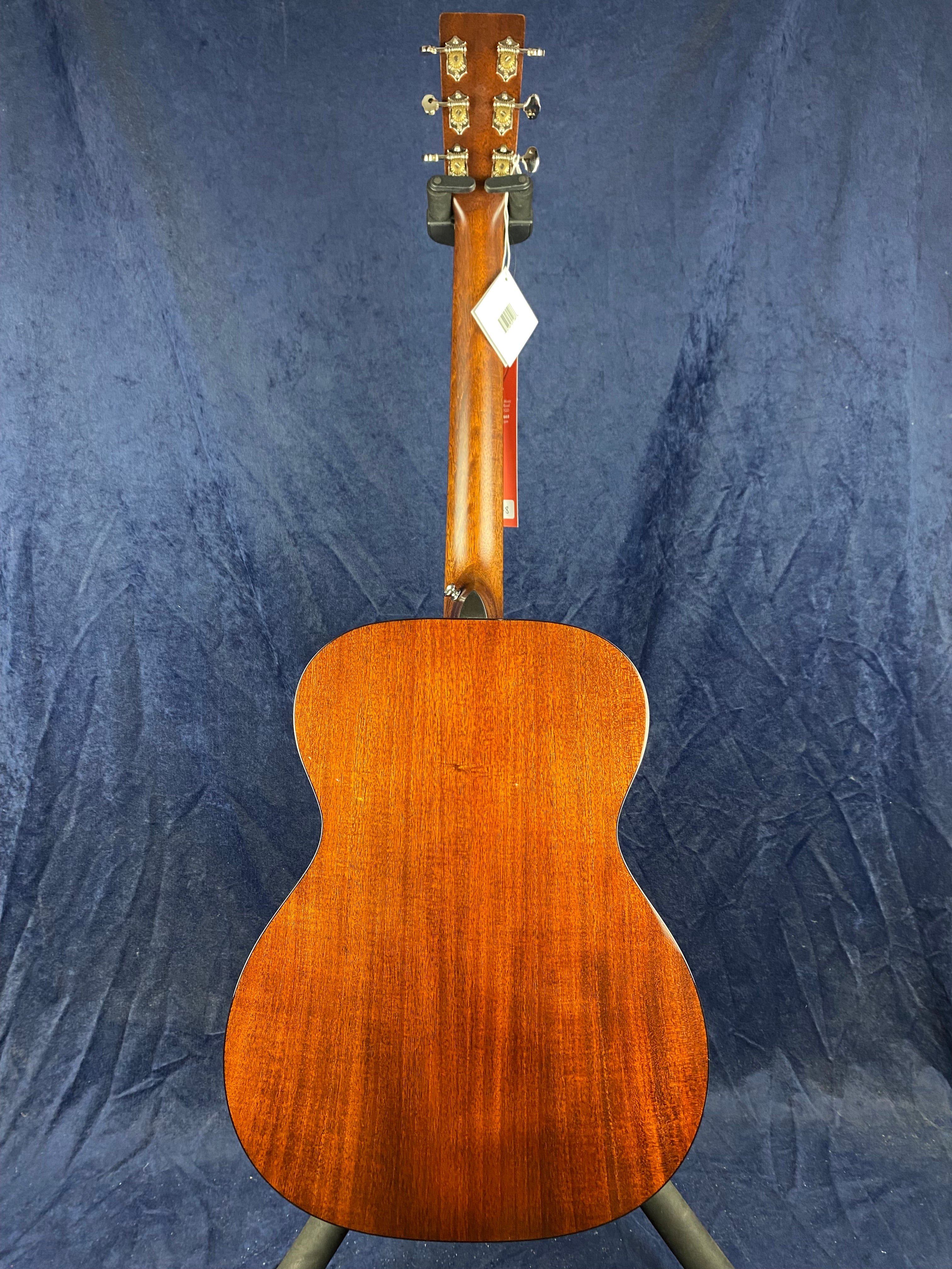 Back view of an acoustic guitar with a smooth, rich brown wood finish, gold tuning pegs, and a white tag hanging from the neck against a dark background.