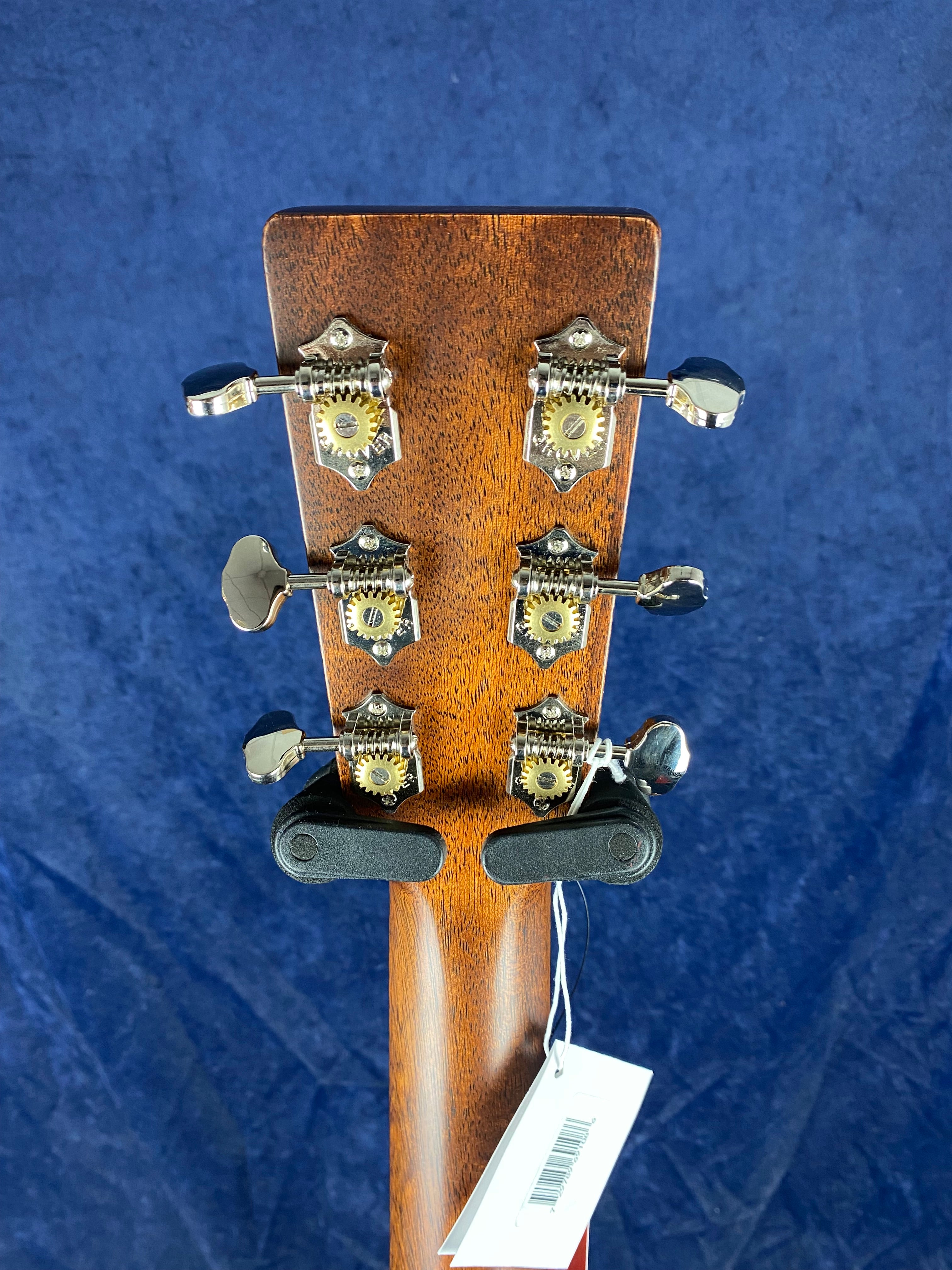 Rear view of an acoustic guitar headstock showing six metal tuning pegs, wood grain finish, and a barcode tag attached with string.