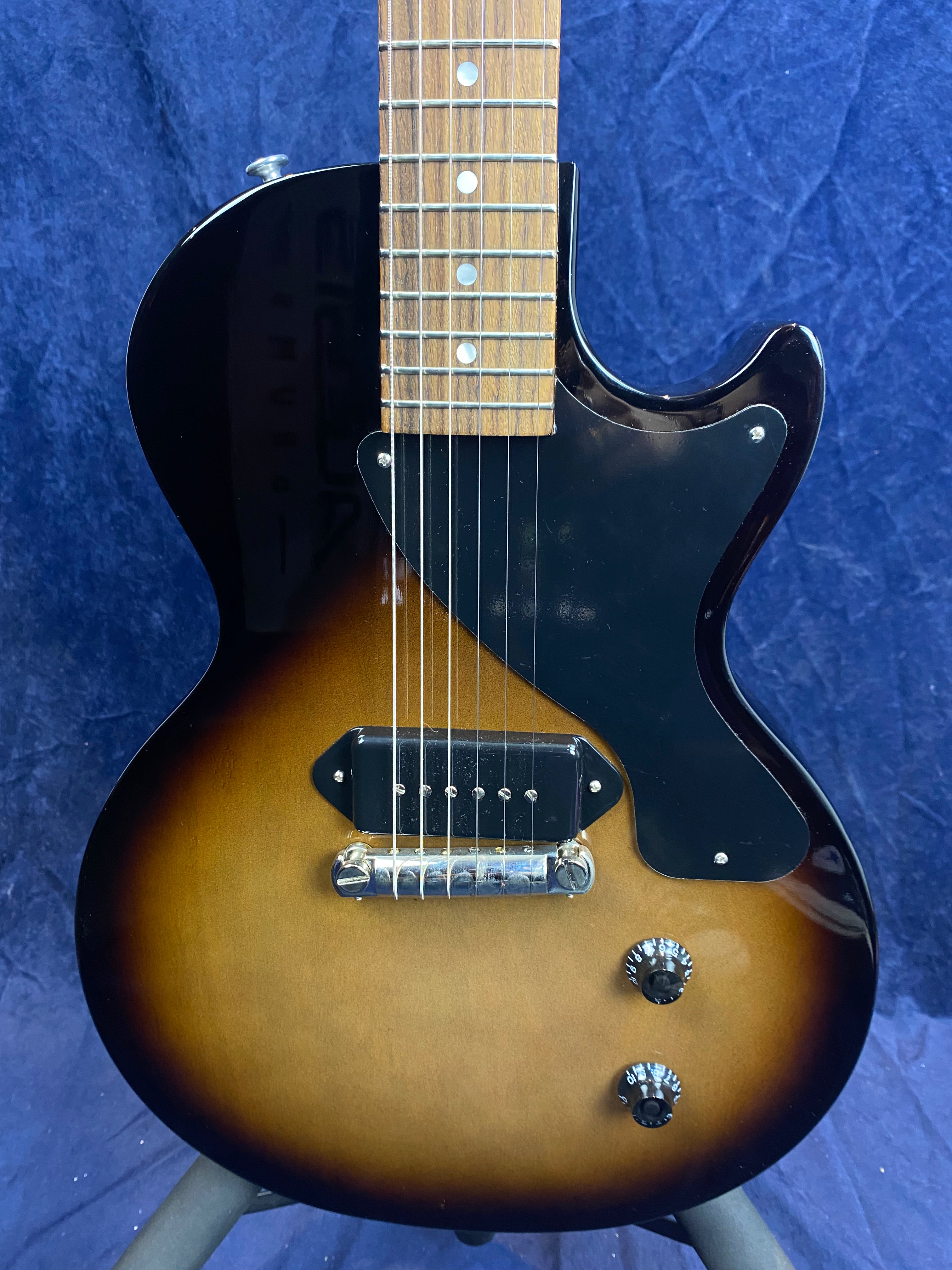 Single-cutaway electric guitar with a sunburst finish, black pickguard, one pickup, two control knobs, metallic bridge, and wood fretboard.