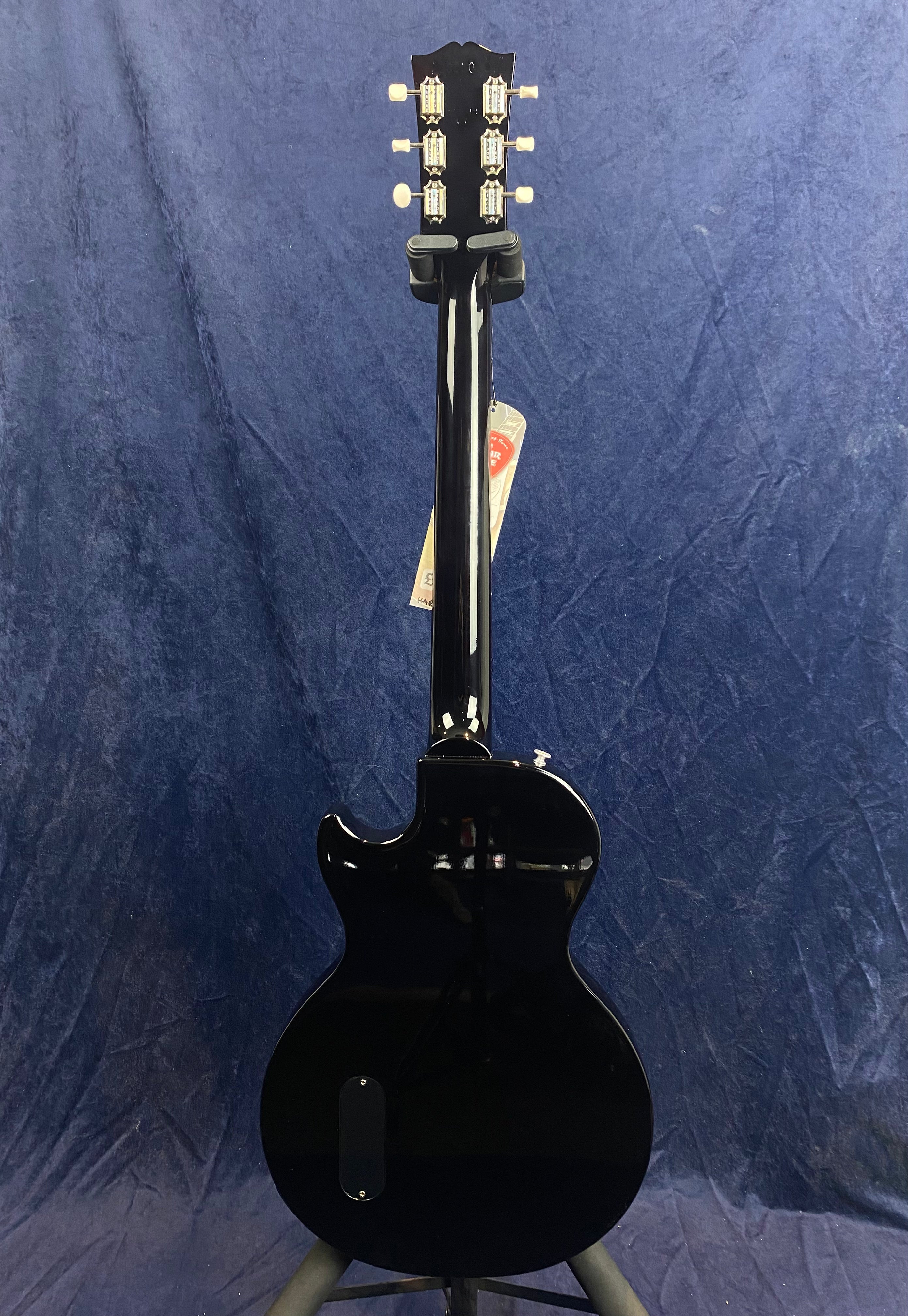 A glossy black electric guitar shown from the back, with six tuning pegs and a single cutaway body, standing against a dark blue background.