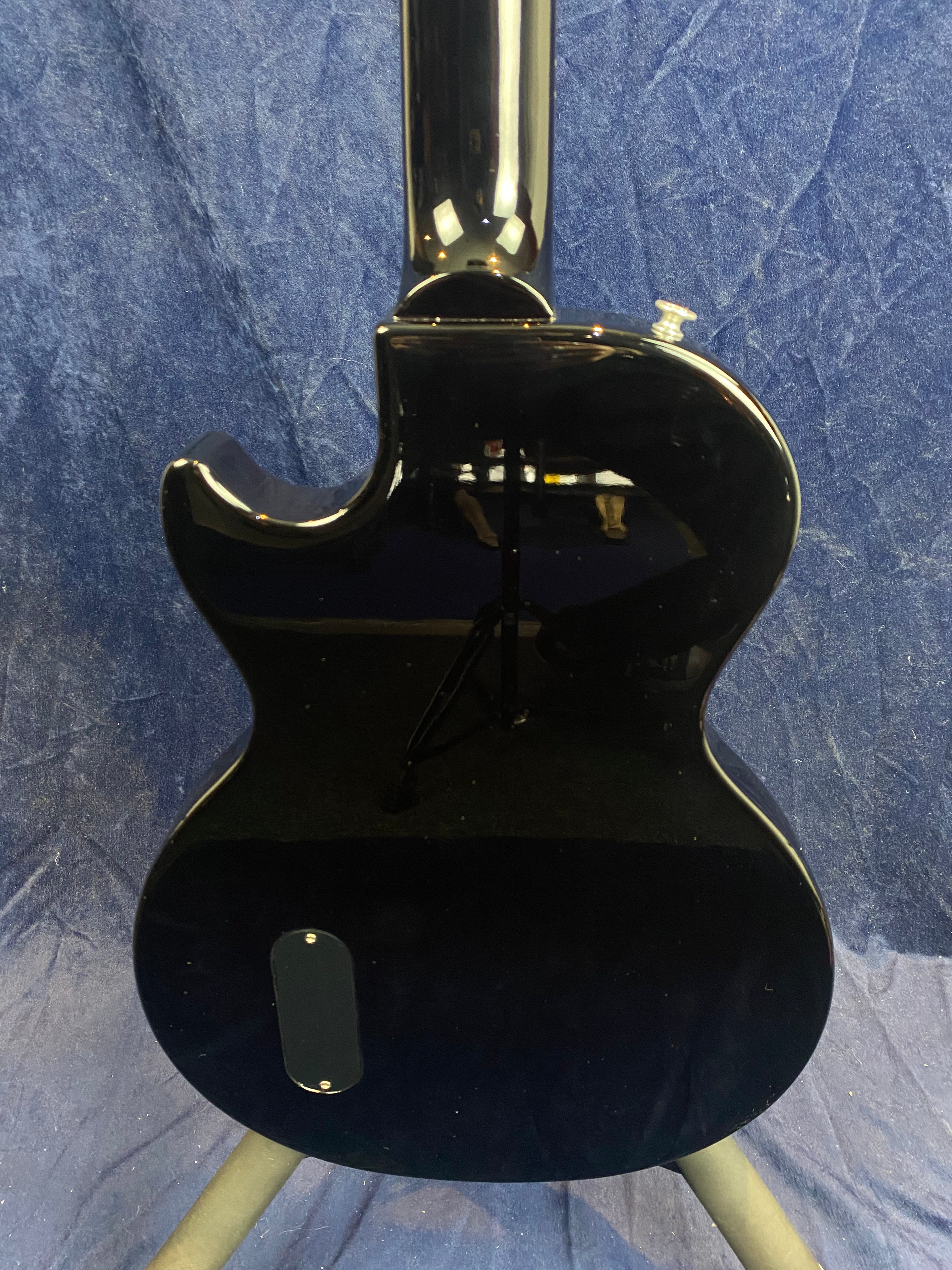Glossy black electric guitar shown from the back, single cutaway shape, oval back plate, standing on a tripod against a blue fabric background.