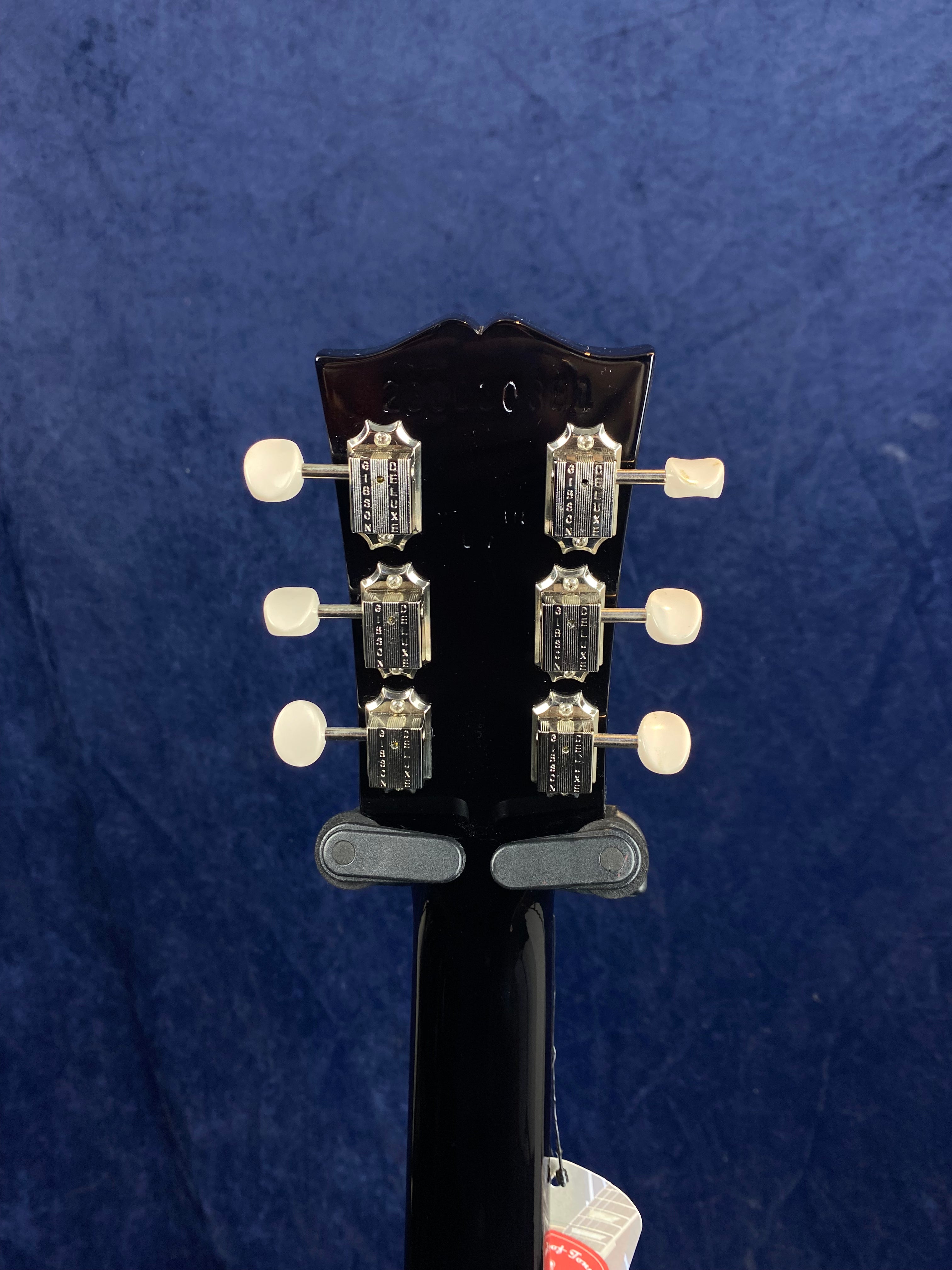 Back of a black electric guitar headstock with six vintage-style Gibson tuners and white tuning pegs, set against a dark blue background.